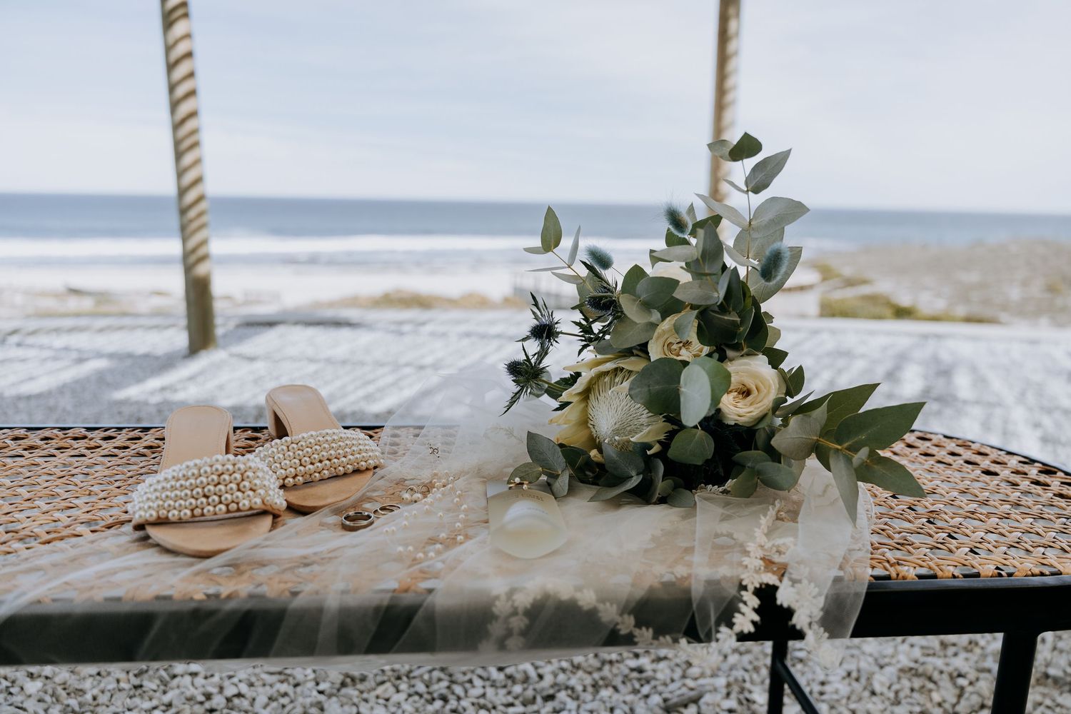 Wedding centerpiece close-up with white flowers and greenery arranged in rope-wrapped vase on a beach table setting.
