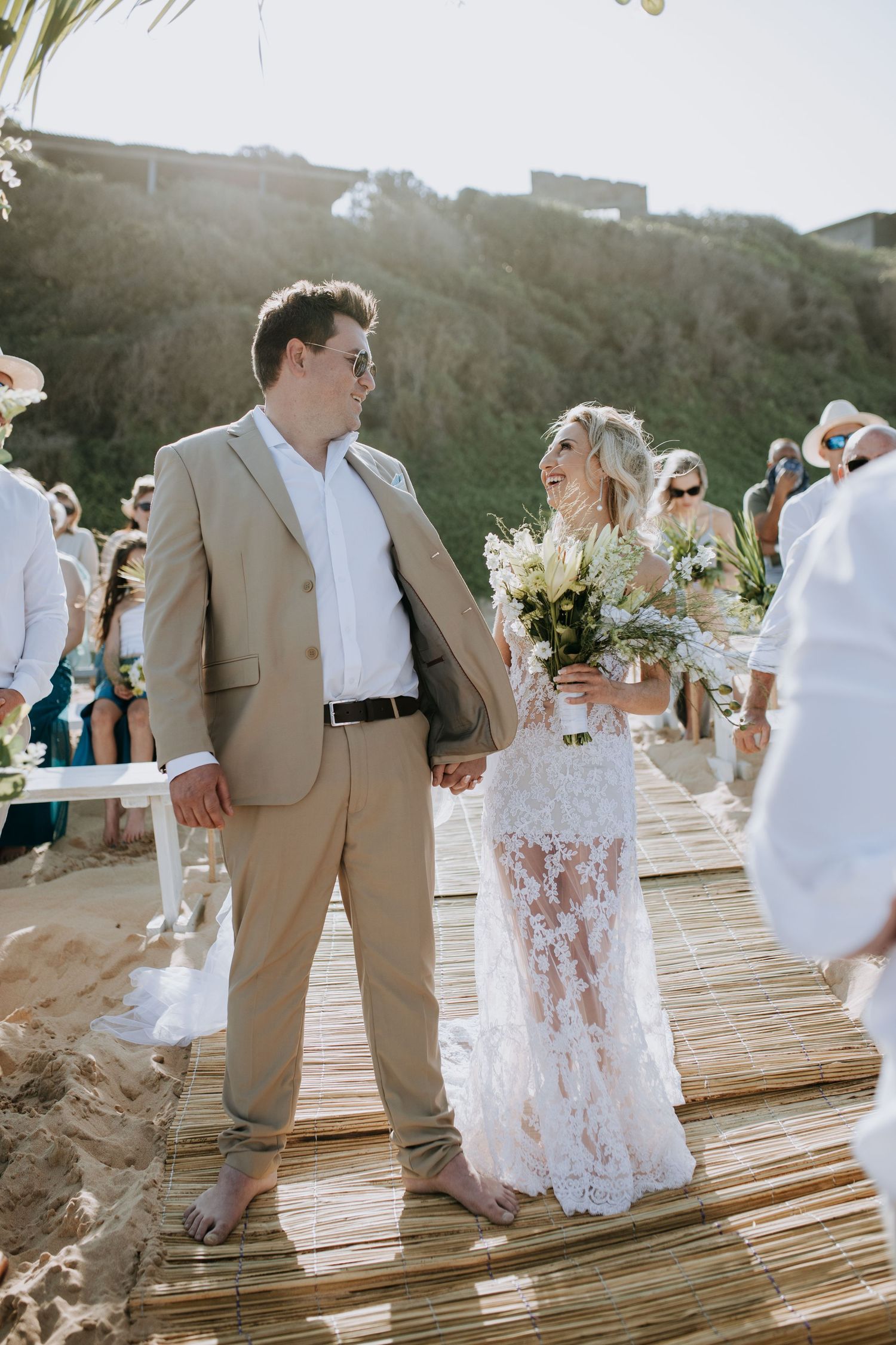 Wedding party members in tan suits and flowing white dresses walk together on a rustic wooden deck during sunset.