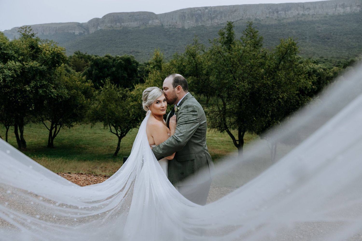 A romantic wedding couple embraces in front of a scenic mountain backdrop while the bride's veil flows in the wind.