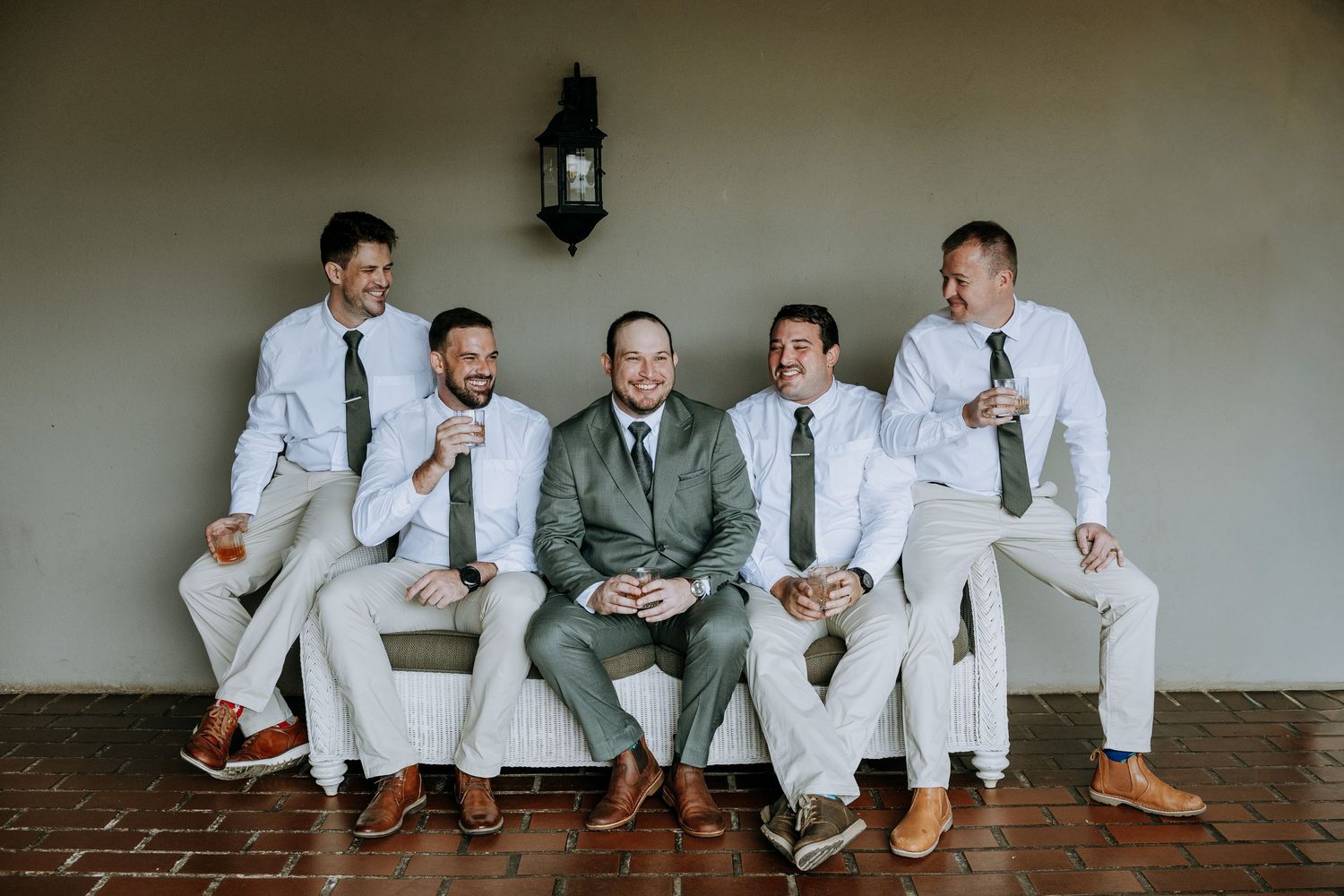 Wedding party in white suits and brown shoes sit casually on a bench in a minimalist indoor setting.