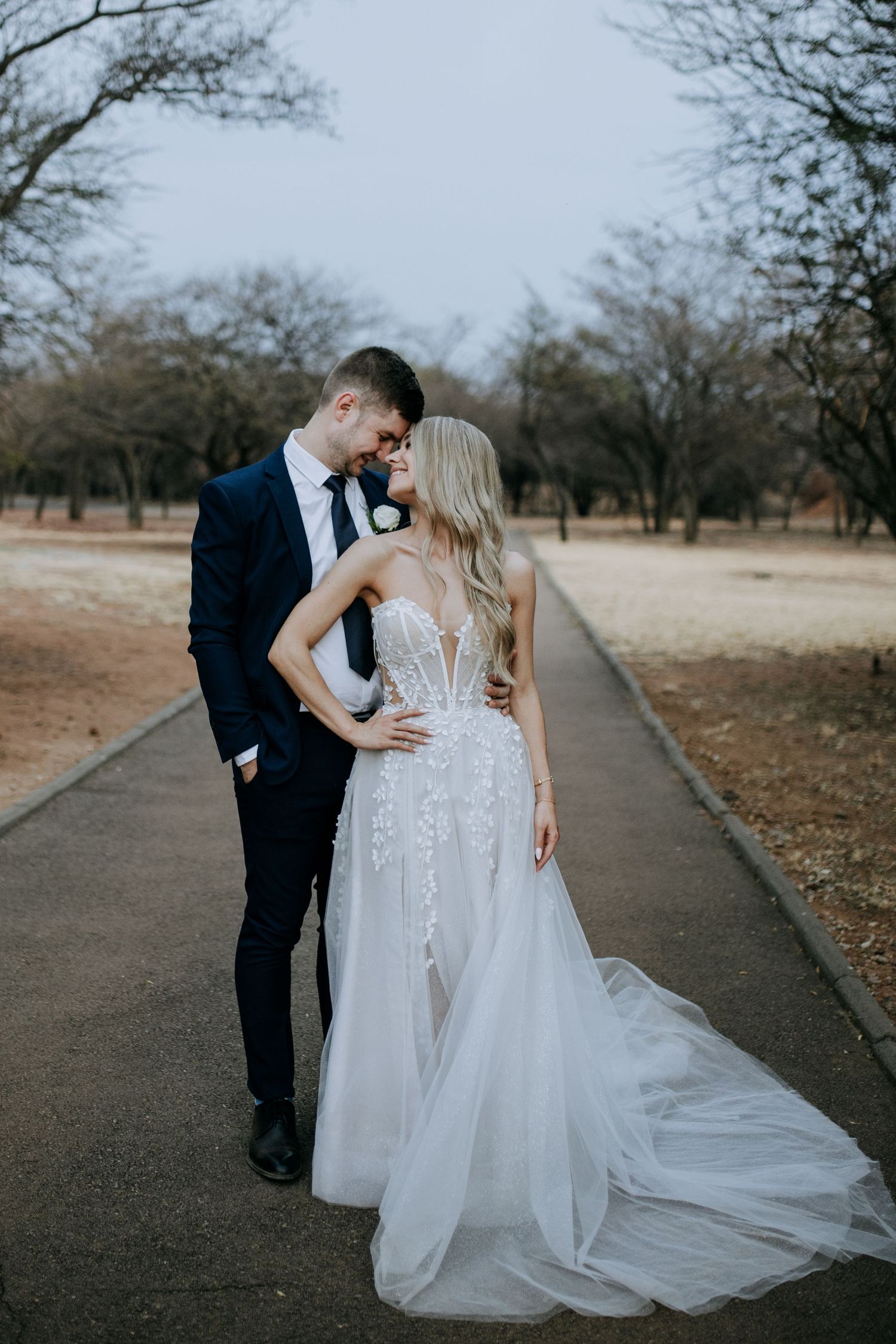 A romantic wedding photo of a couple embracing on a concrete path surrounded by trees.