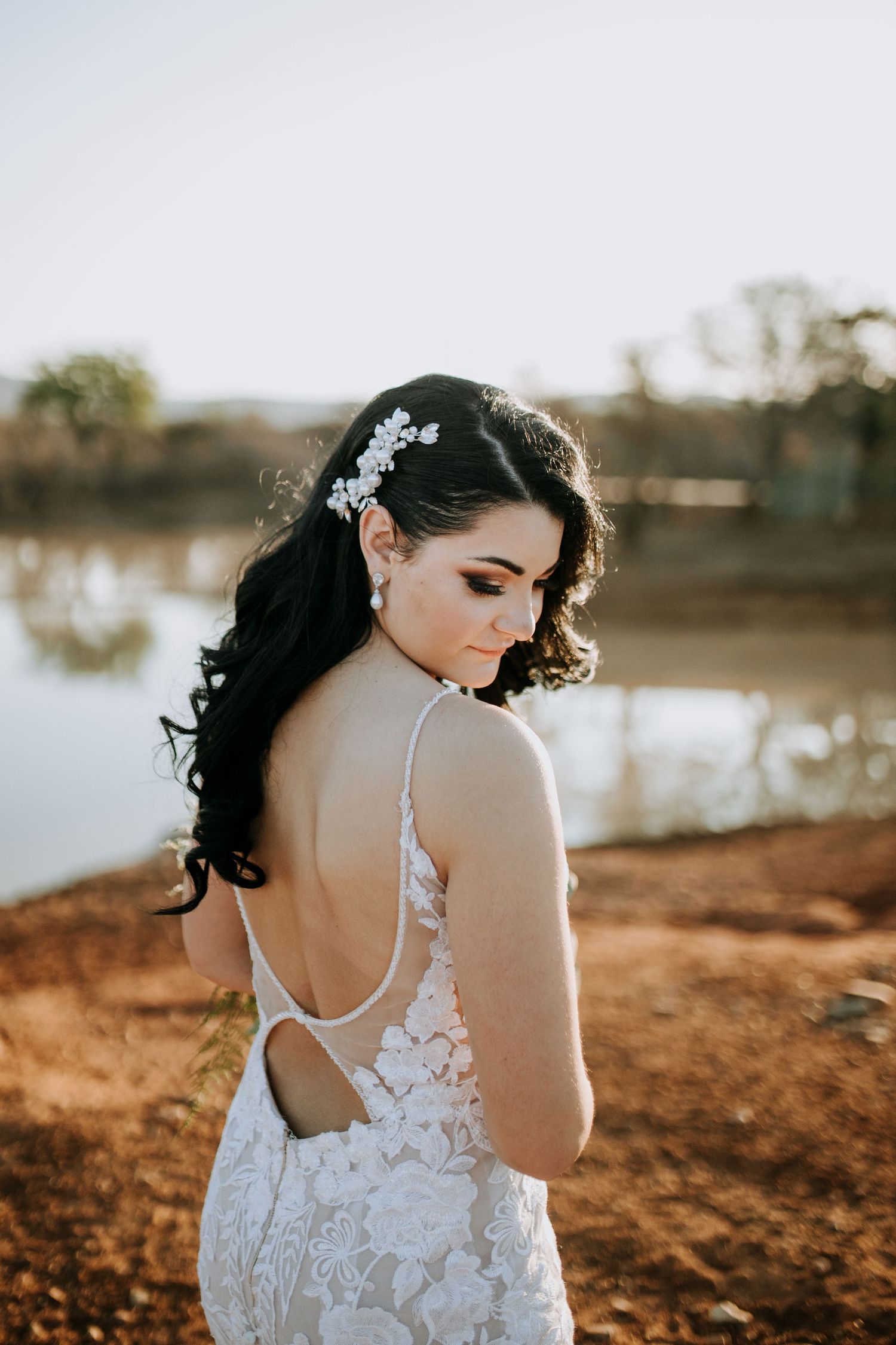 A bride in a lace backless wedding gown poses near a lake at sunset with delicate floral hair accessories.