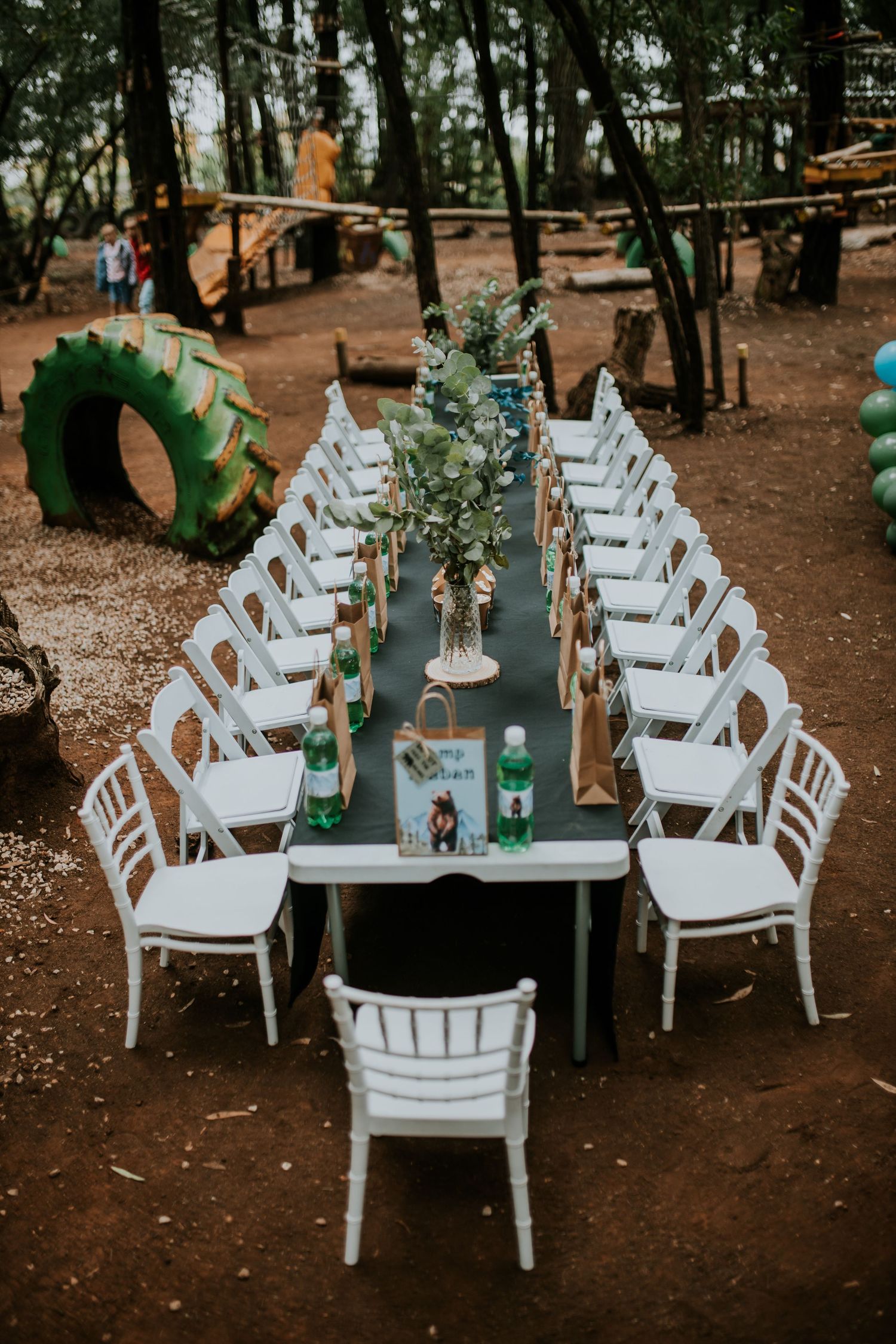 Rustic outdoor dining table with white chairs and greenery decorations set up in a wooded area with tire playground elements.