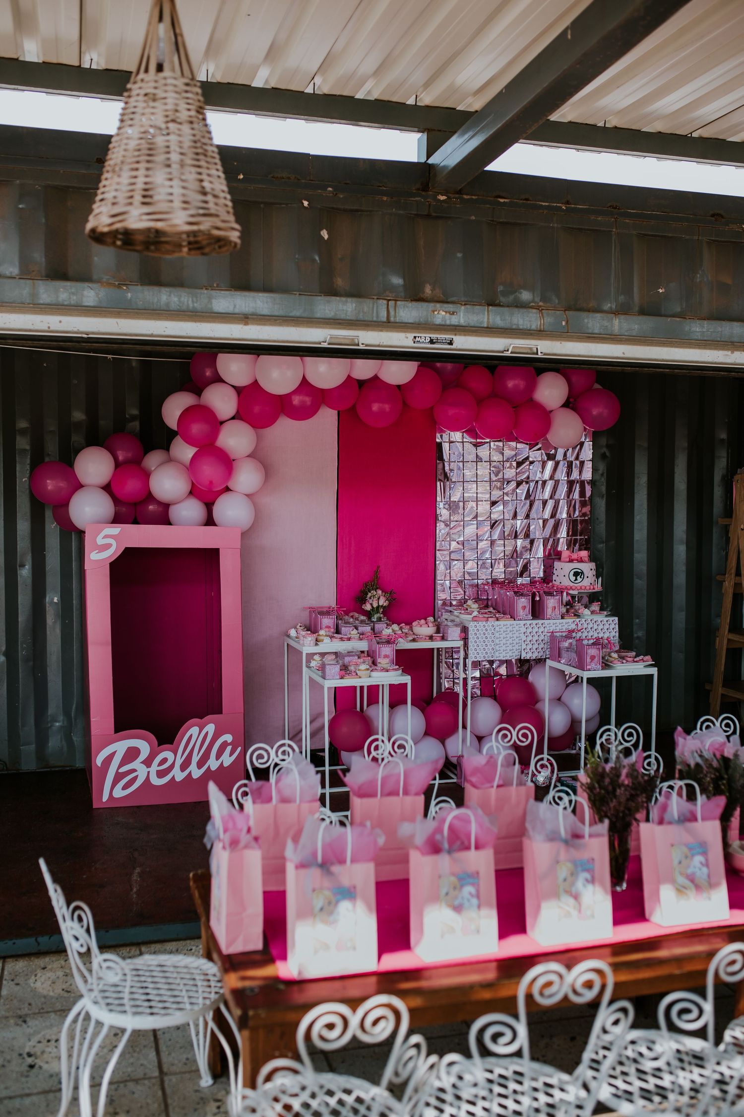 Party setup with pink and white balloon decorations, white chairs, and gift bags arranged in an elegant indoor venue.