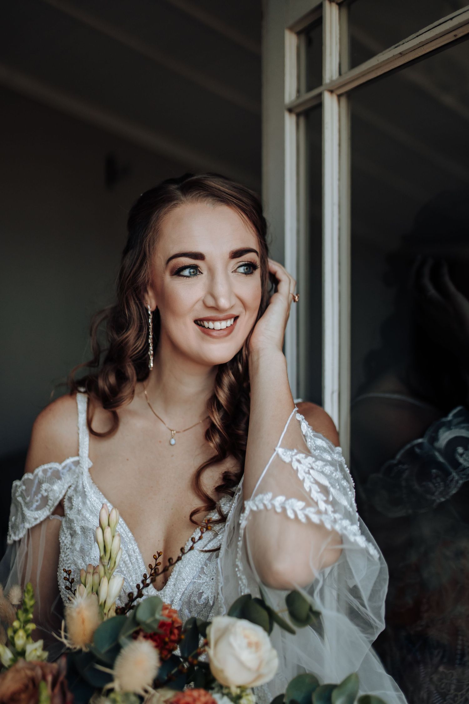 A bride in a lace wedding gown smiles near a window while holding a bouquet of pastel roses.
