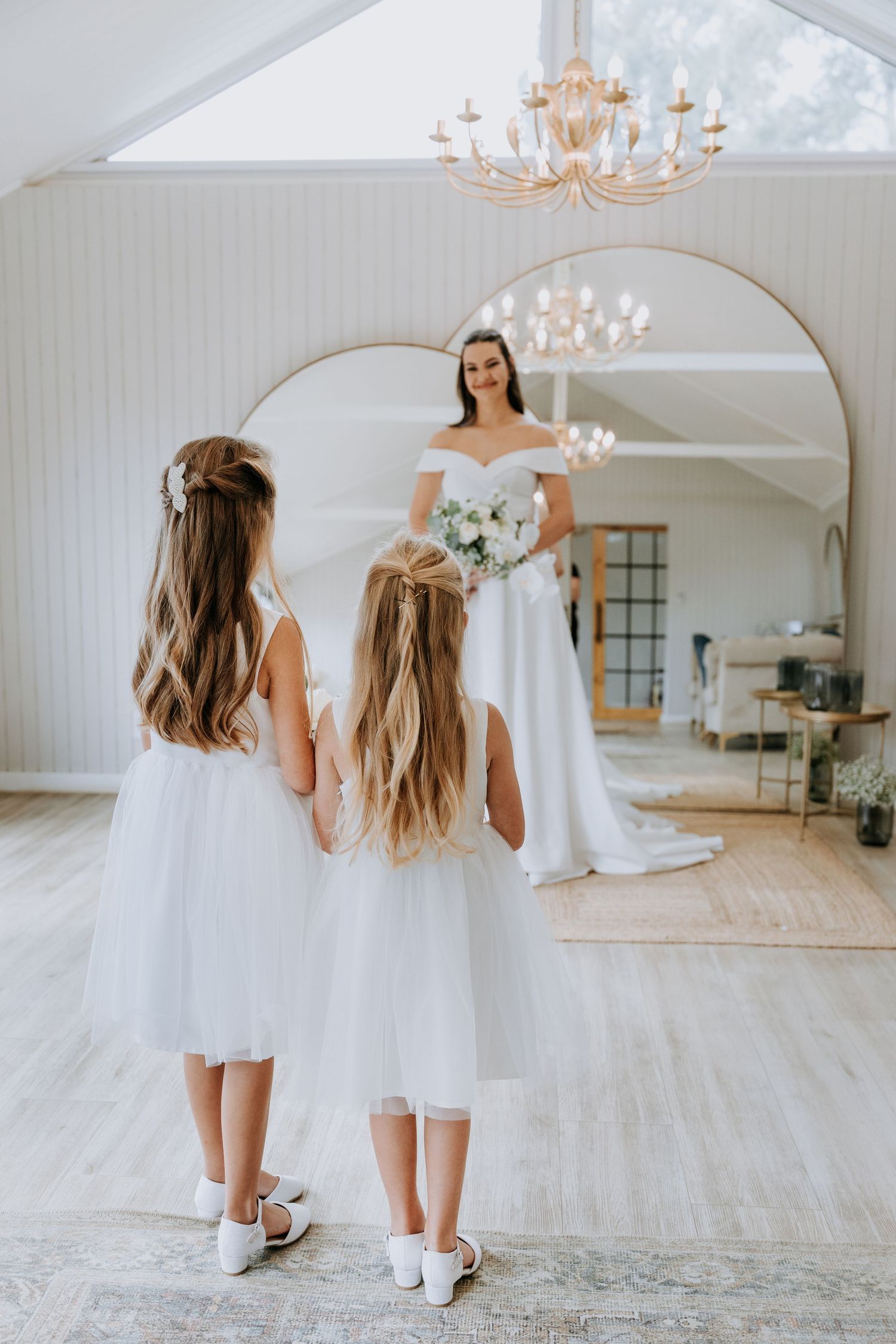 Two flower girls in white dresses look at bride in wedding gown standing before large mirror in bright room with chandelier.