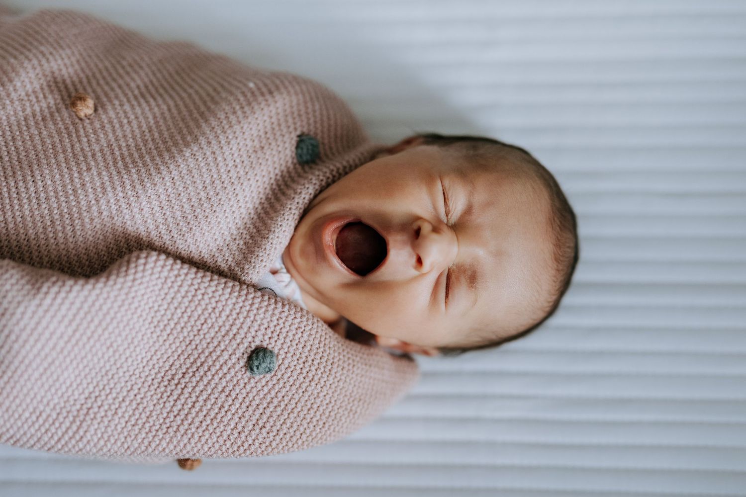 Newborn baby yawning while wrapped in a cozy pink knit swaddle blanket, lying on a white surface.