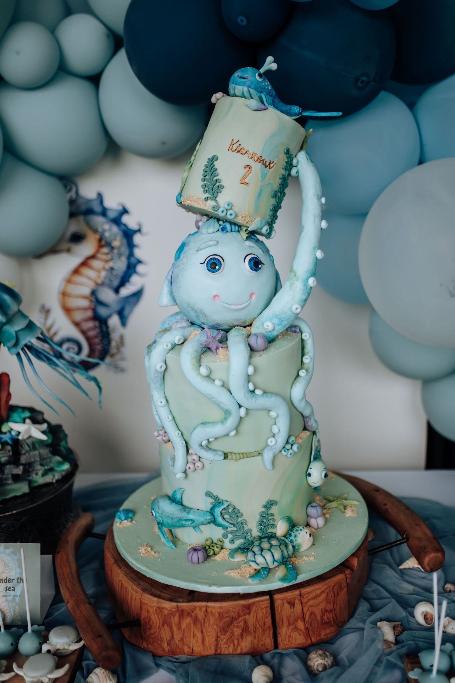 A whimsical under-the-sea themed cake with blue and green fondant octopus design on a rustic wooden cake stand shows multiple angles.