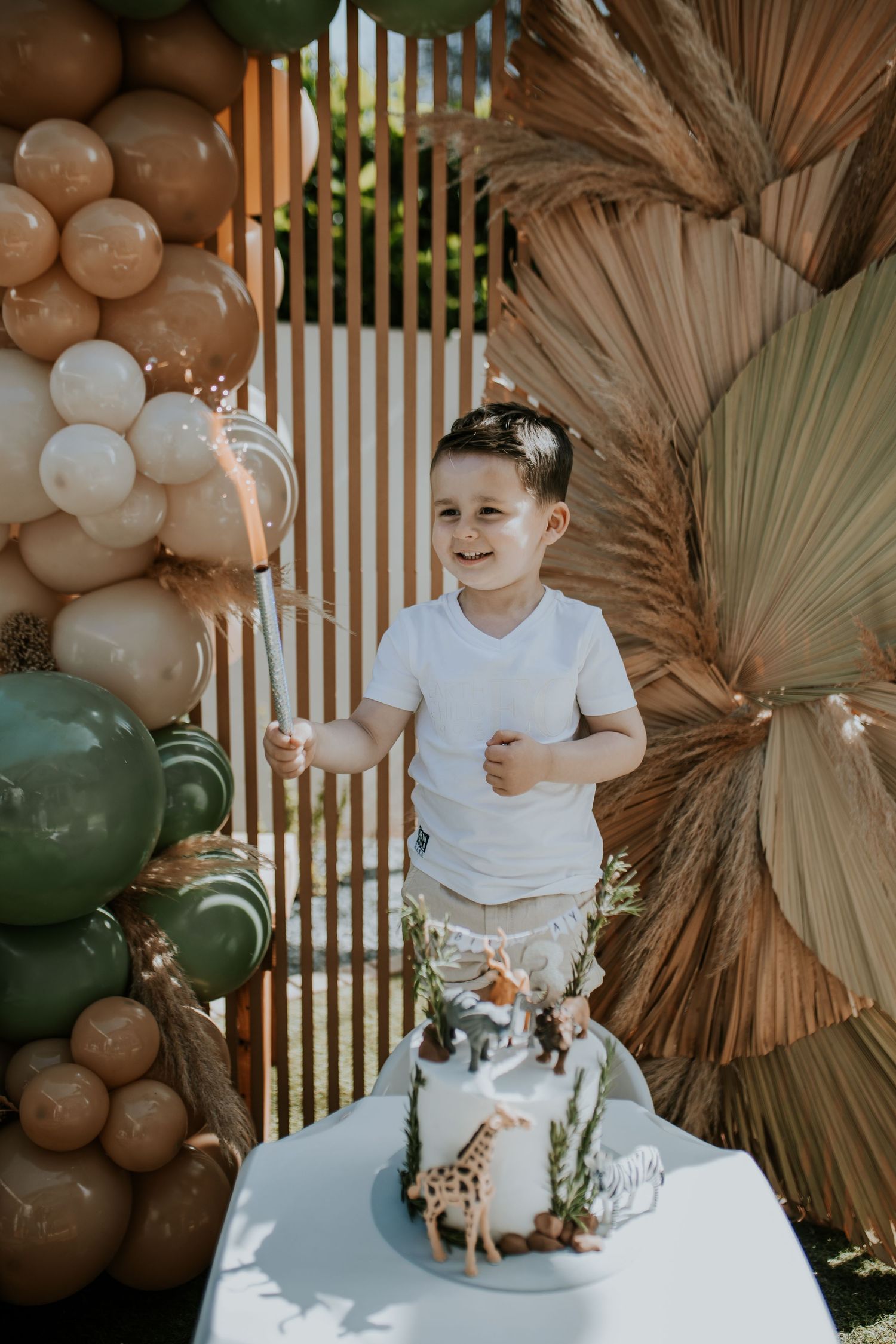 Modern boho party decor with dried palm leaves, white grid backdrop and neutral balloon garland in earth tones.