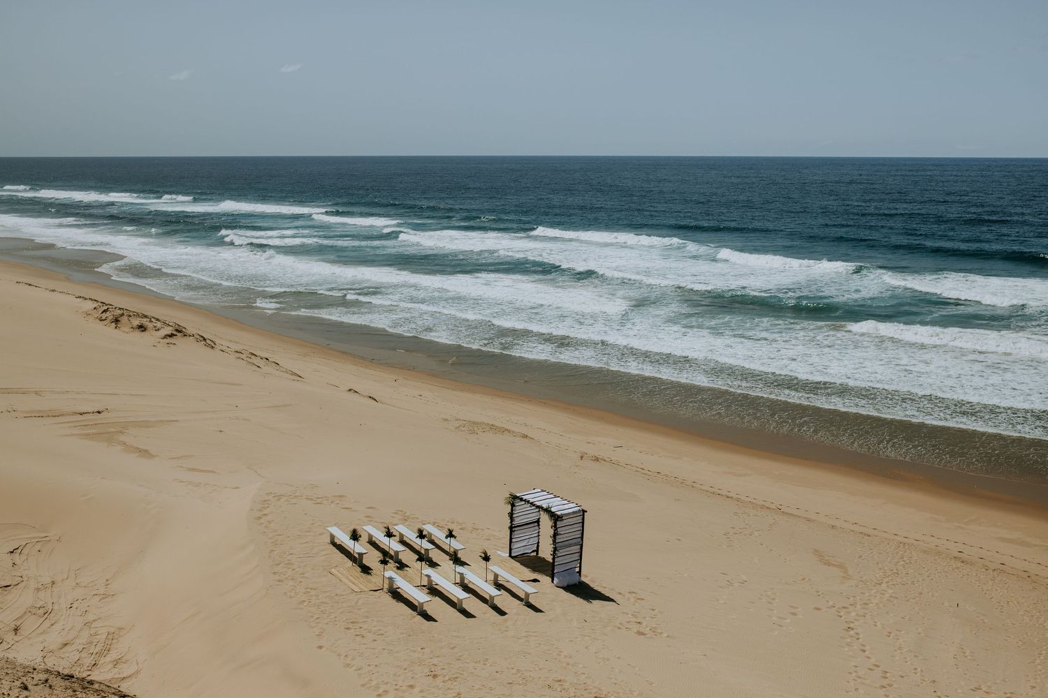 A beach wedding setup with white chairs and arch facing the ocean waves on an empty sandy beach.