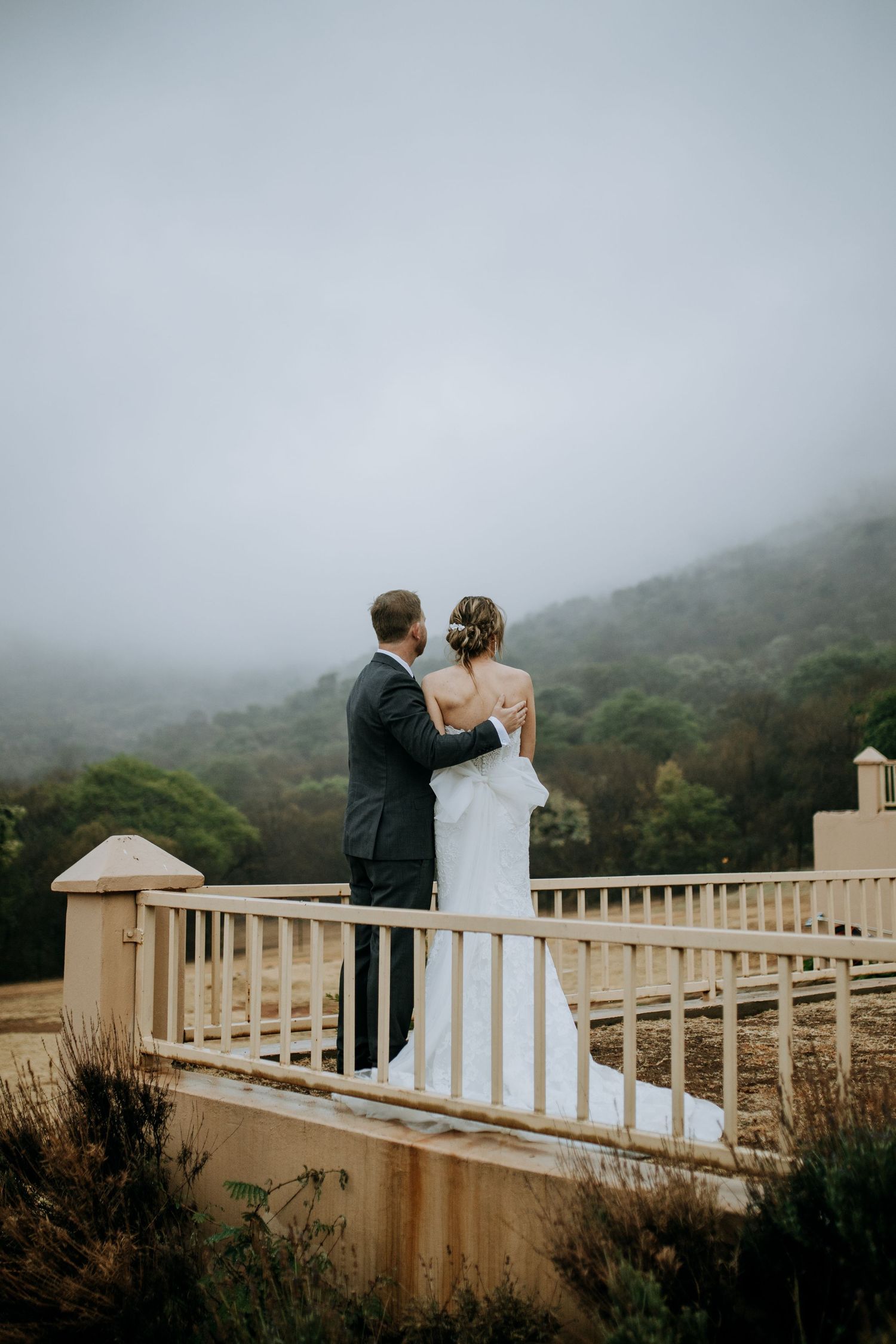 A couple sharing a romantic moment on a misty coastal overlook with cream-colored railings and foggy mountains in view.
