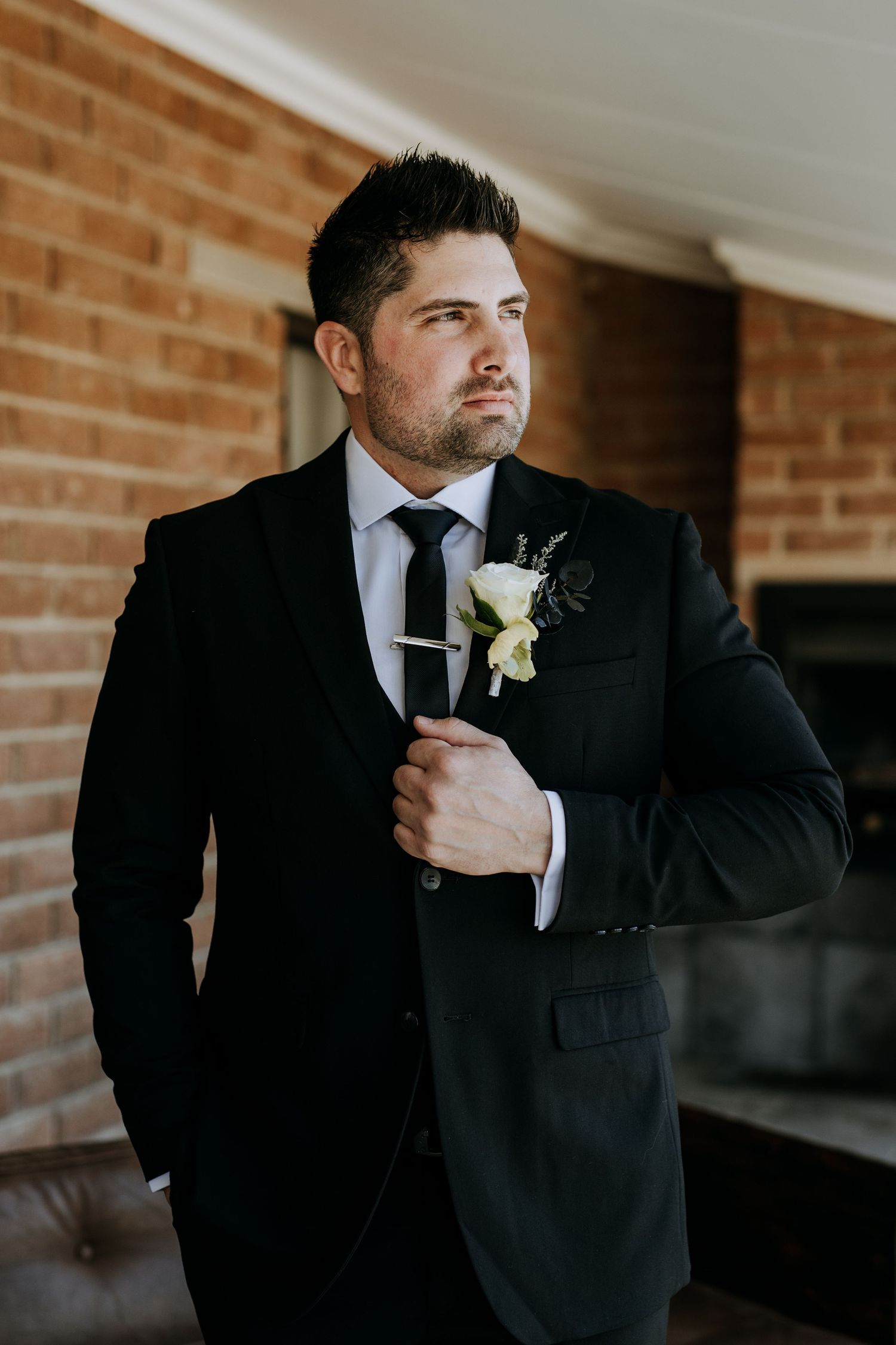 A groom in a black suit and tie straightens his boutonniere against a brick wall in a series of candid photos.