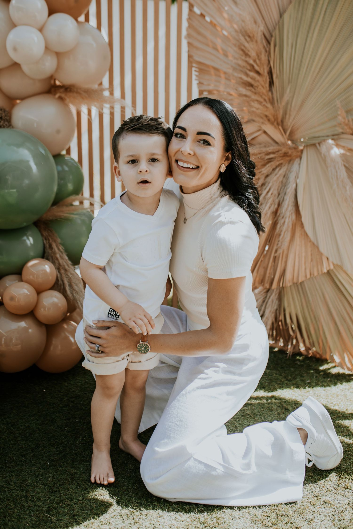 A heartwarming outdoor photoshoot featuring a white-dressed duo posing against dried palm leaves and balloon decorations.