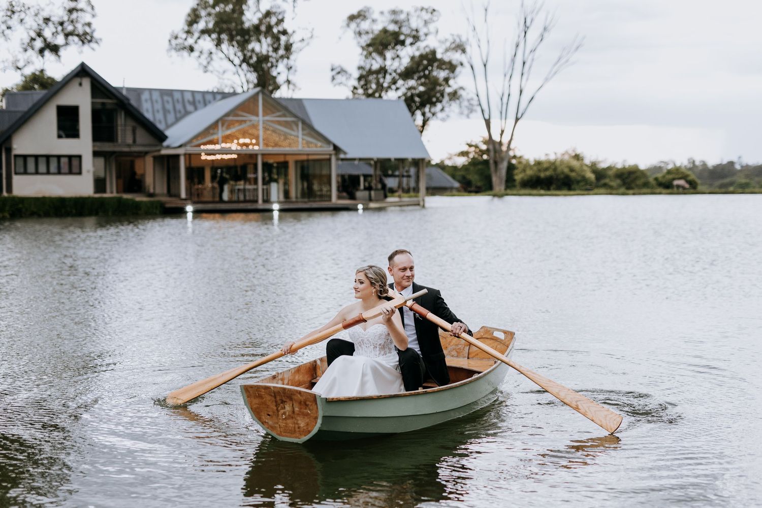 A wooden rowboat glides across calm waters with two occupants paddling together on a misty day.