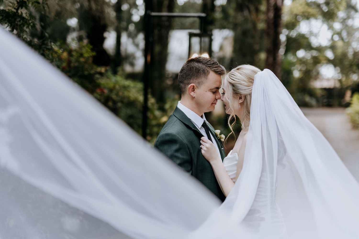 A romantic wedding couple embraces outdoors in a garden setting with soft natural lighting and flowing white veil.