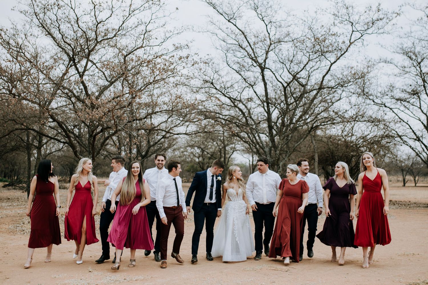 Wedding party in burgundy dresses and black suits poses together in a desert landscape with bare trees.
