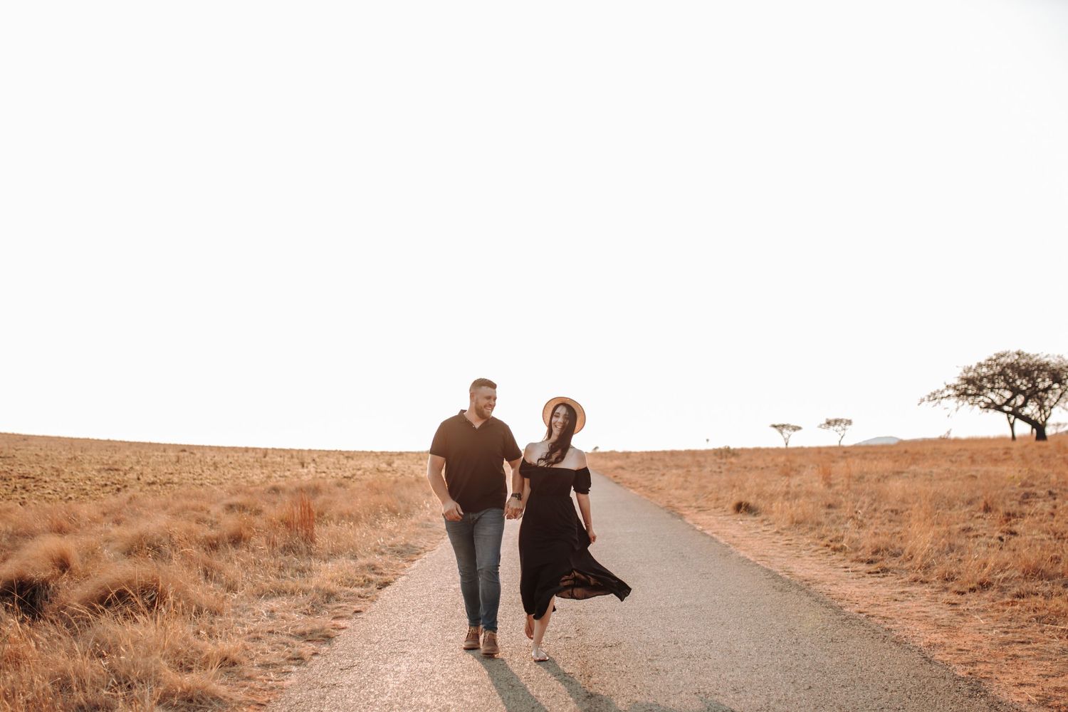 Two people walk down a sunlit road through a scenic desert landscape at sunset.