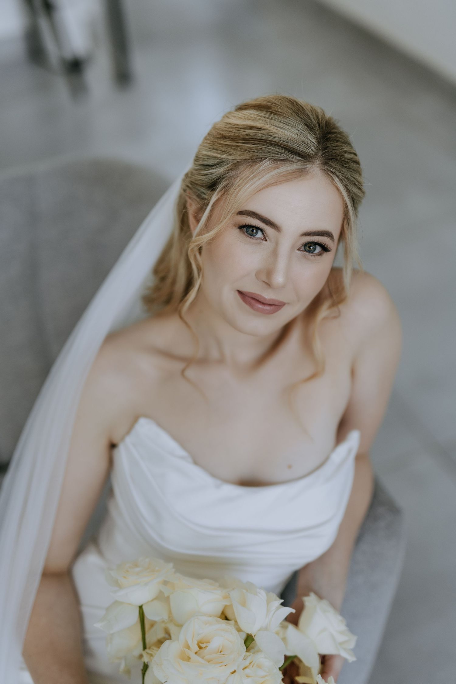 Bridal portrait featuring a white strapless gown and cream-colored rose bouquet in an elegant indoor setting.