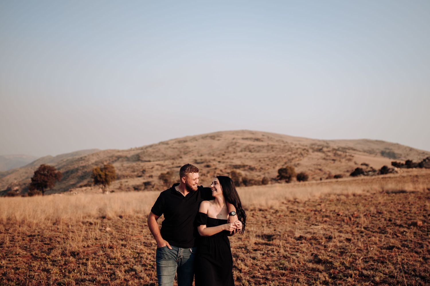 A couple embraces in warm golden light on a rugged hilltop at sunset with rolling hills in the background.