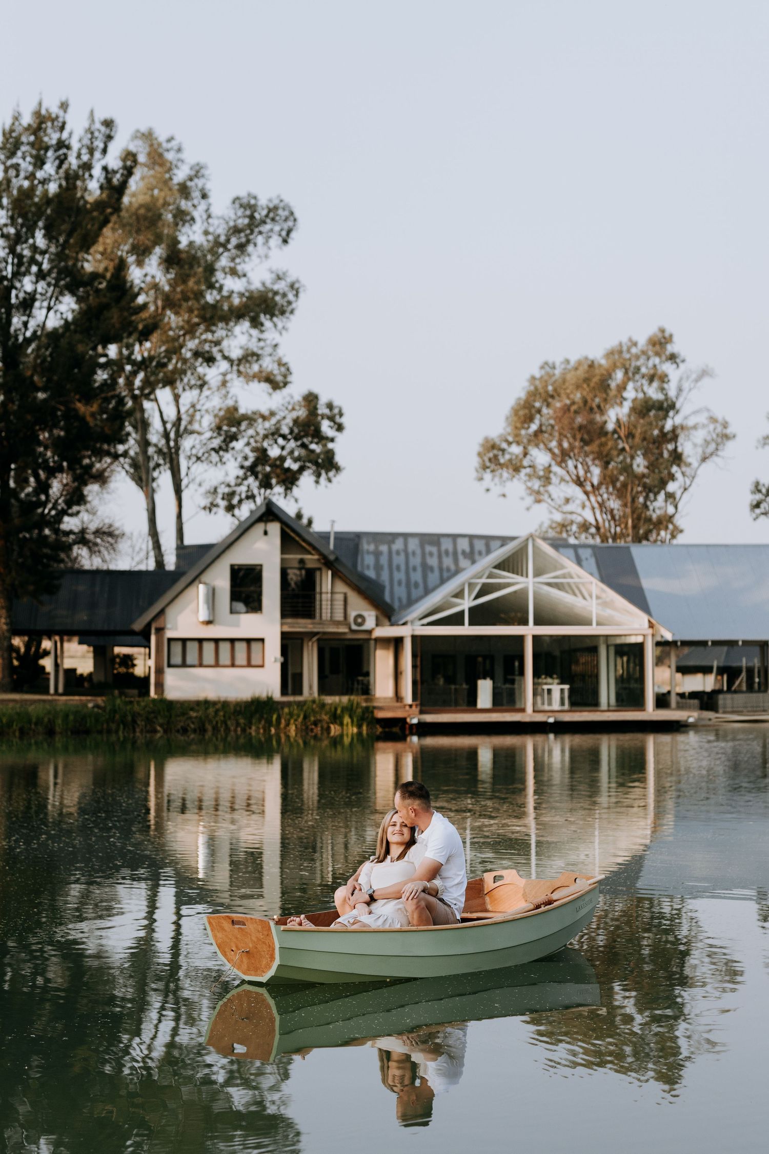 Couple relaxes in green wooden boat at lakeside resort with modern waterfront cottages on a tranquil afternoon.