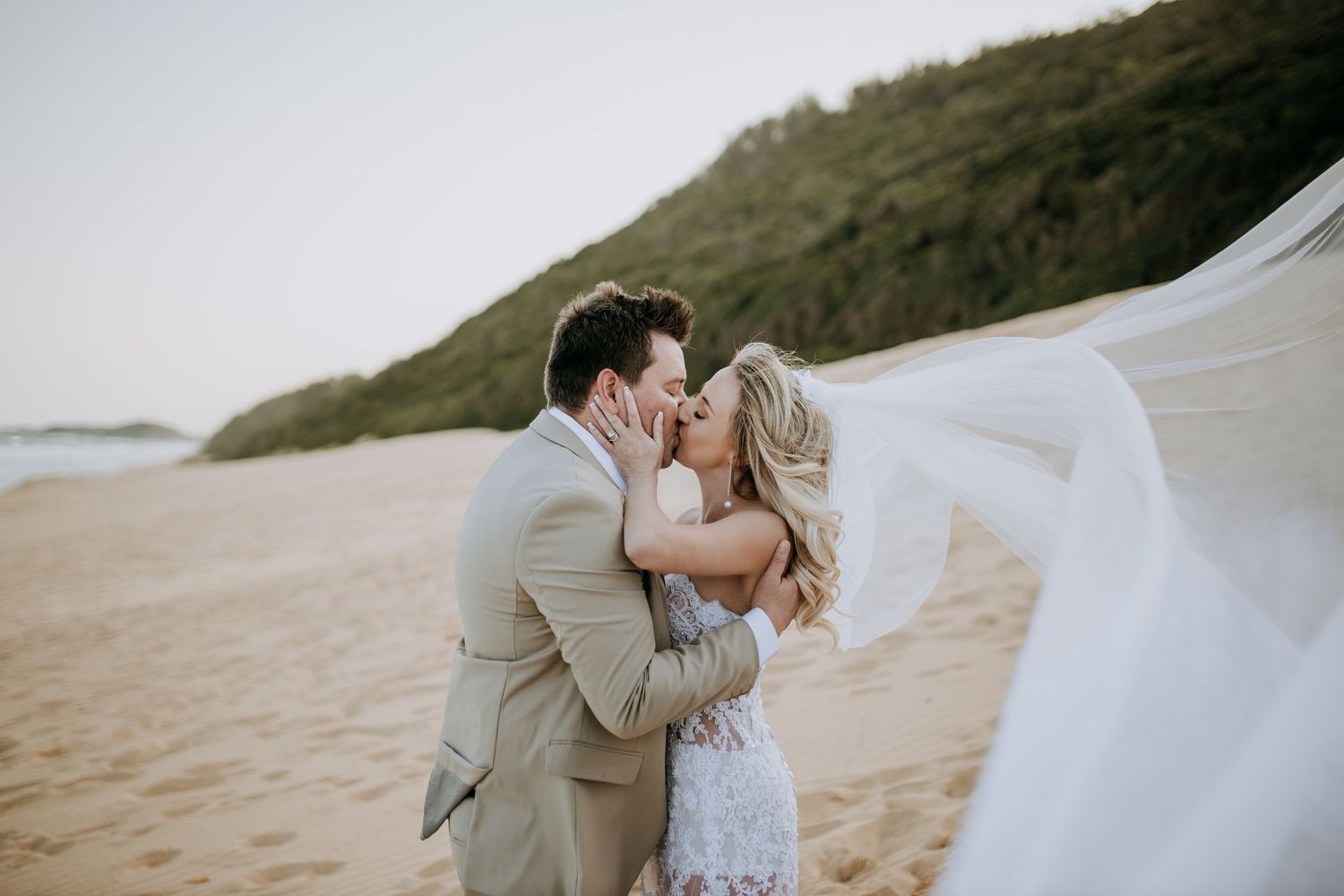 A romantic couple shares an intimate moment on a sandy beach during sunset wearing formal attire.