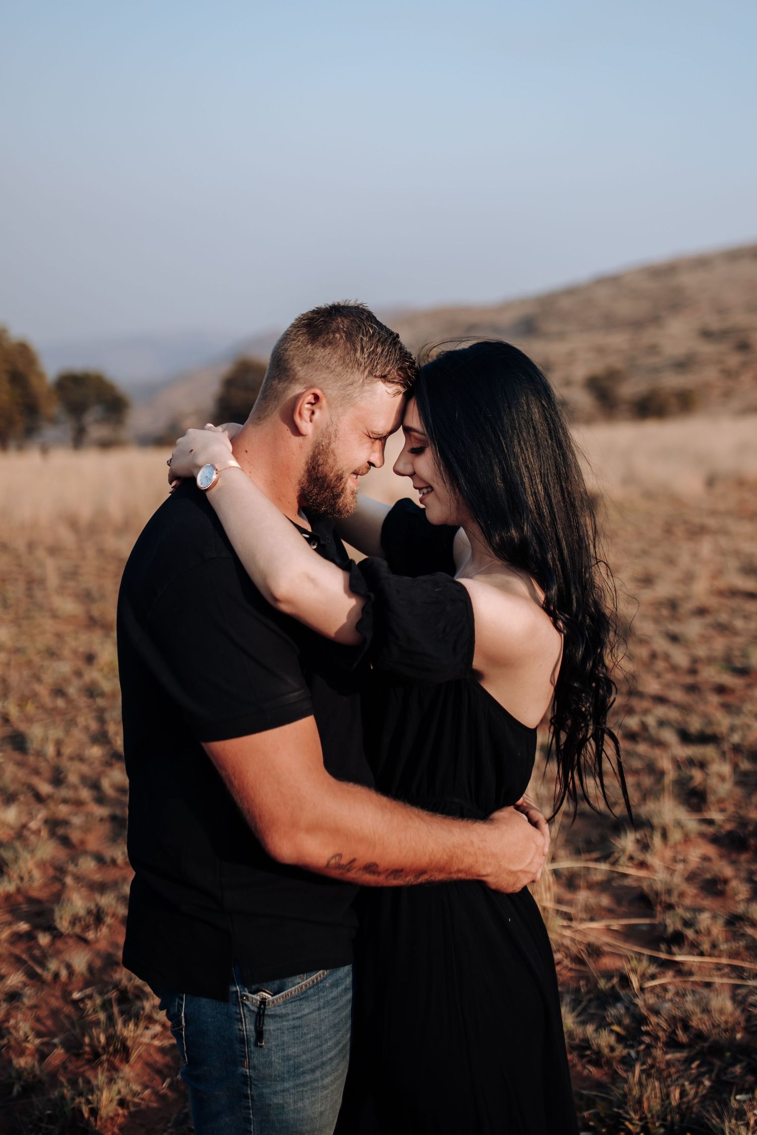 Romantic couple in black outfits share intimate moments while embracing in a dry desert landscape at sunset.