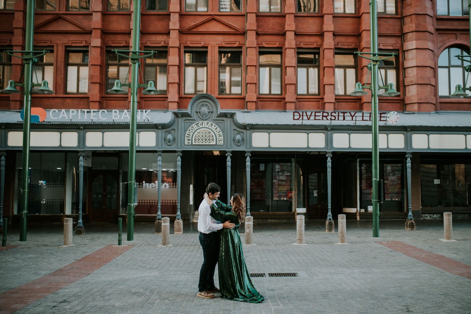 A couple in formal attire embraces in front of a historic brick building with green pillars and arched windows.