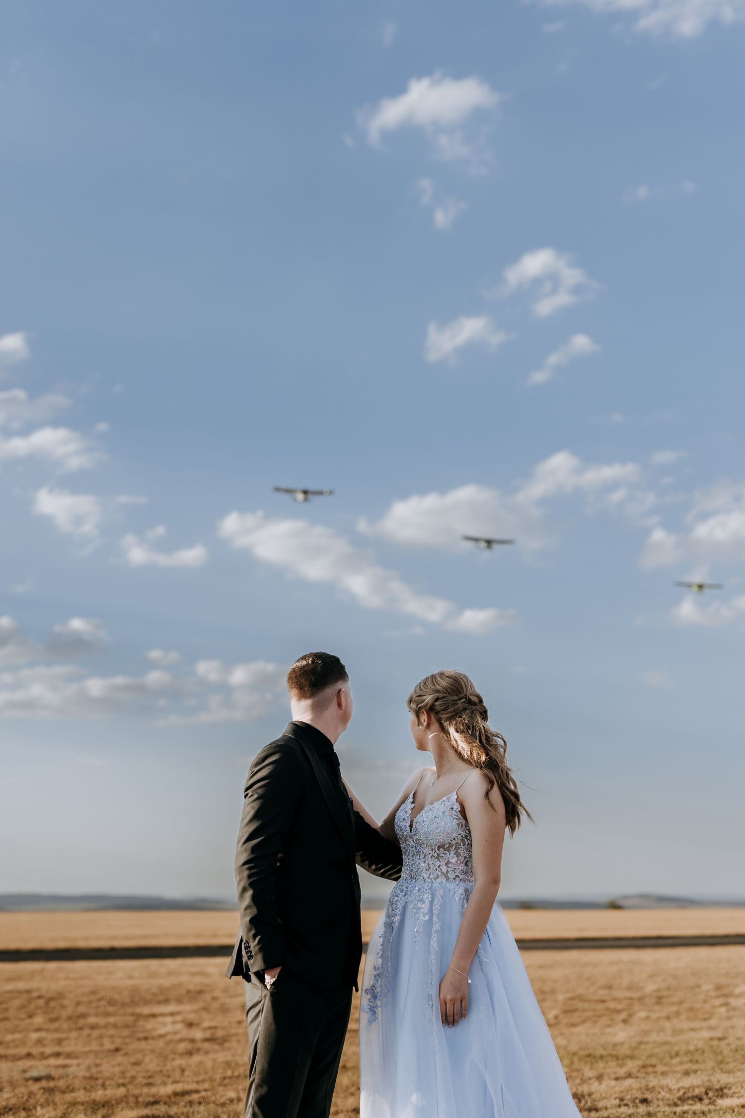 A couple stands together in a golden wheat field under a blue sky with small planes flying overhead.