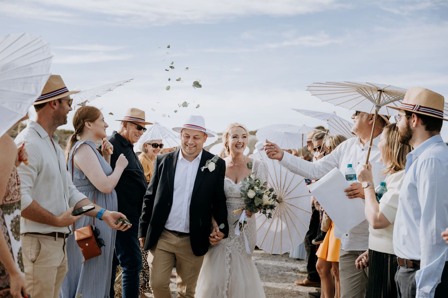 Wedding guests in light summer attire socialize and celebrate on a beach under a bright sky.