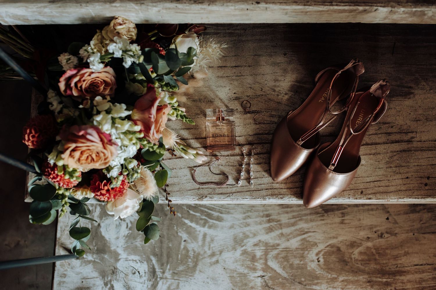 Wedding details including floral arrangements, rings, signage, shoes and delicate decor displayed on rustic wooden surfaces.