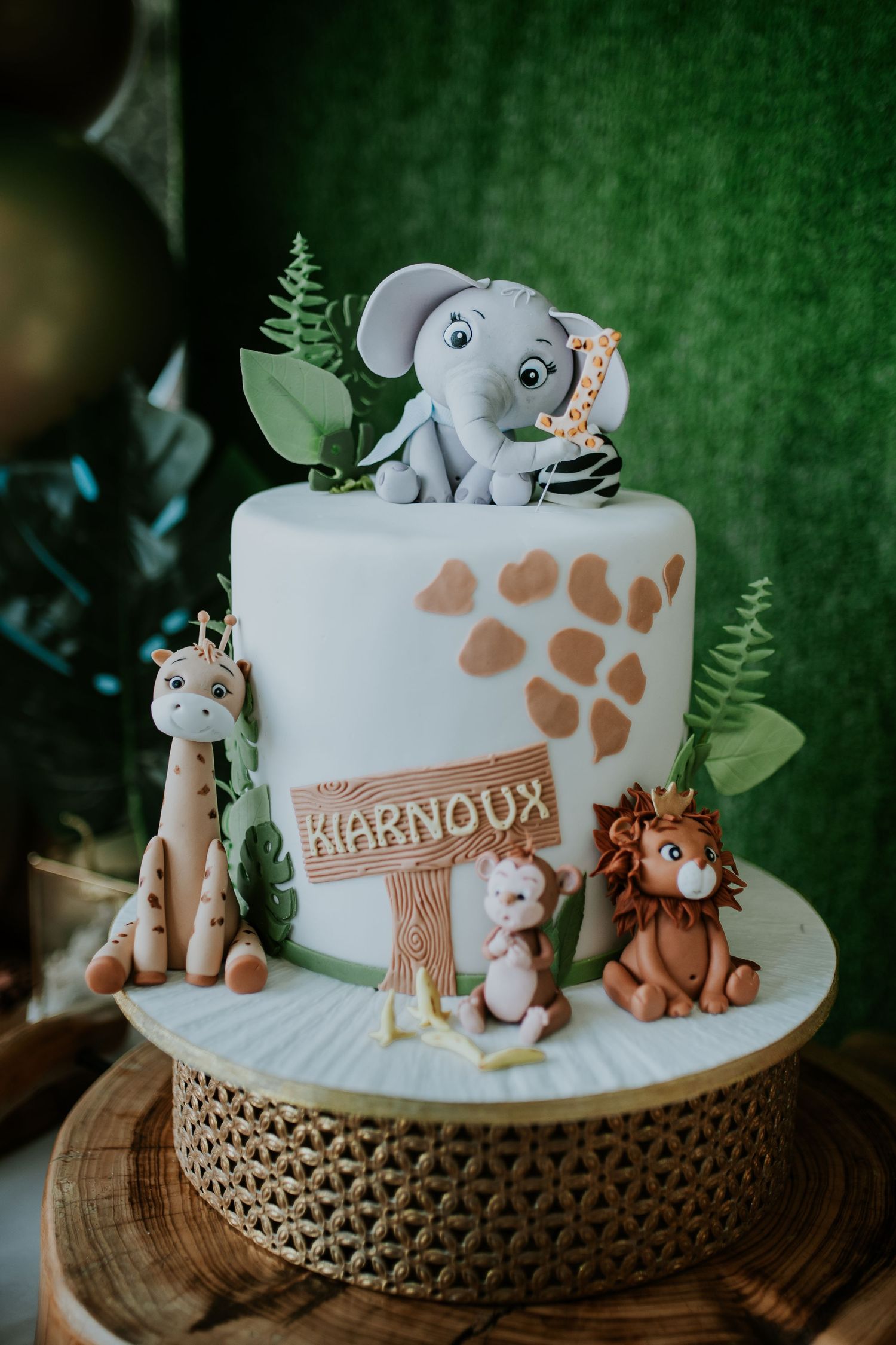 A safari-themed birthday cake decorated with fondant animals and leaves on a wooden cake stand features elegant details.