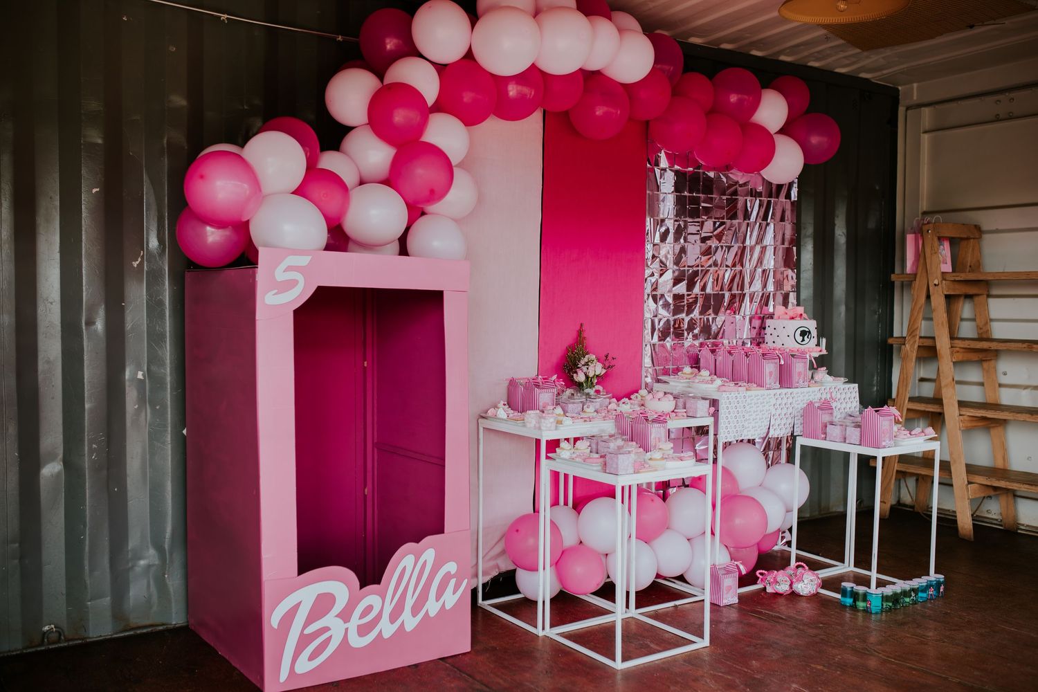 Pink and white balloon decorations create a festive atmosphere in an indoor party space with banners and displays.