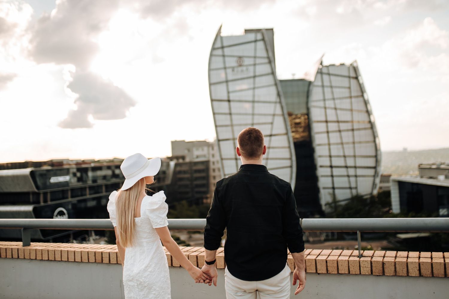 A couple sits on a brick wall overlooking a scenic view during sunset with modern buildings in the background.