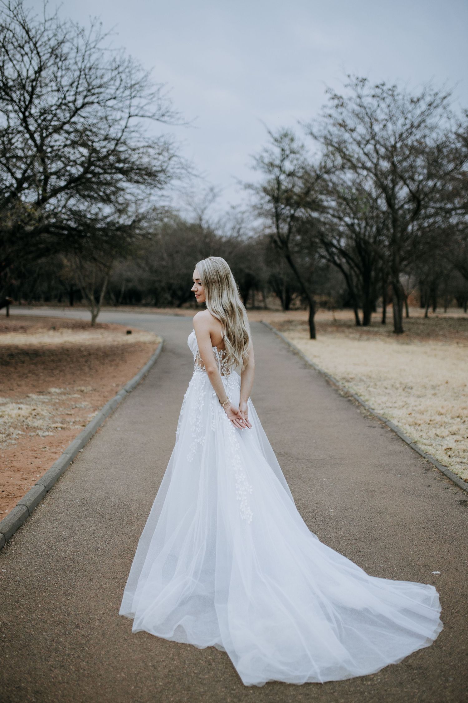 A bride in a flowing white gown stands on a pathway surrounded by bare winter trees.