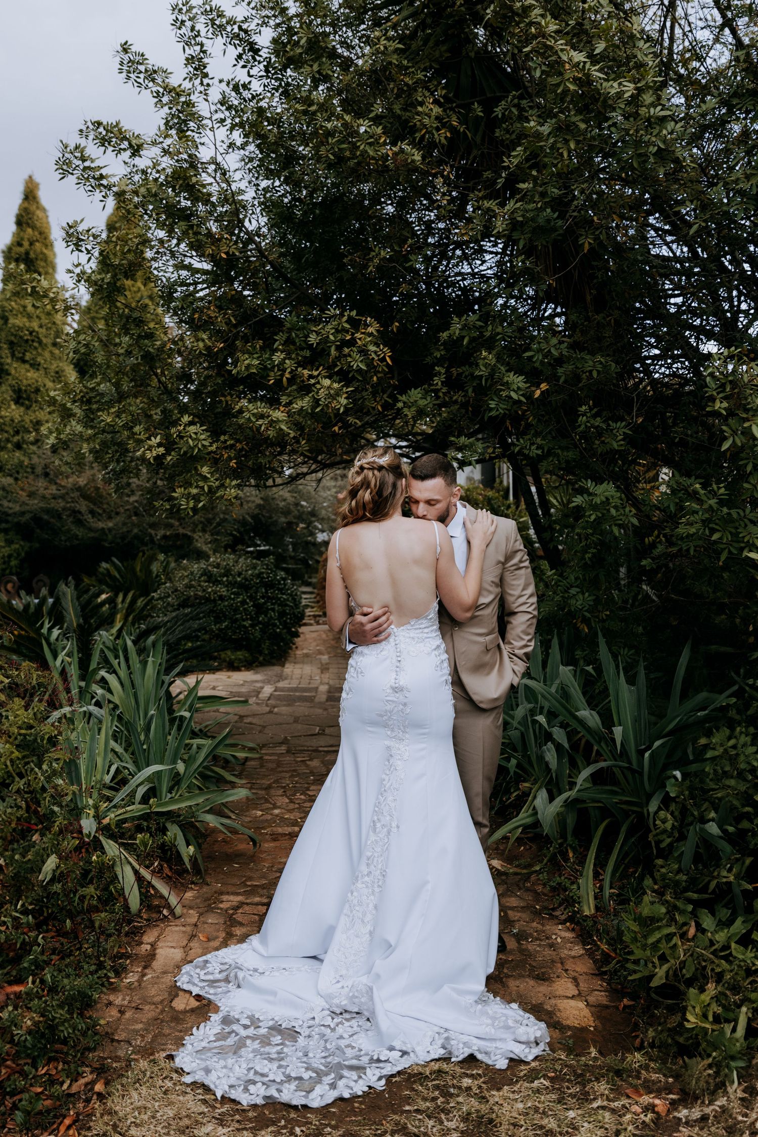 A couple in wedding attire walk along a garden path surrounded by lush greenery and agave plants.