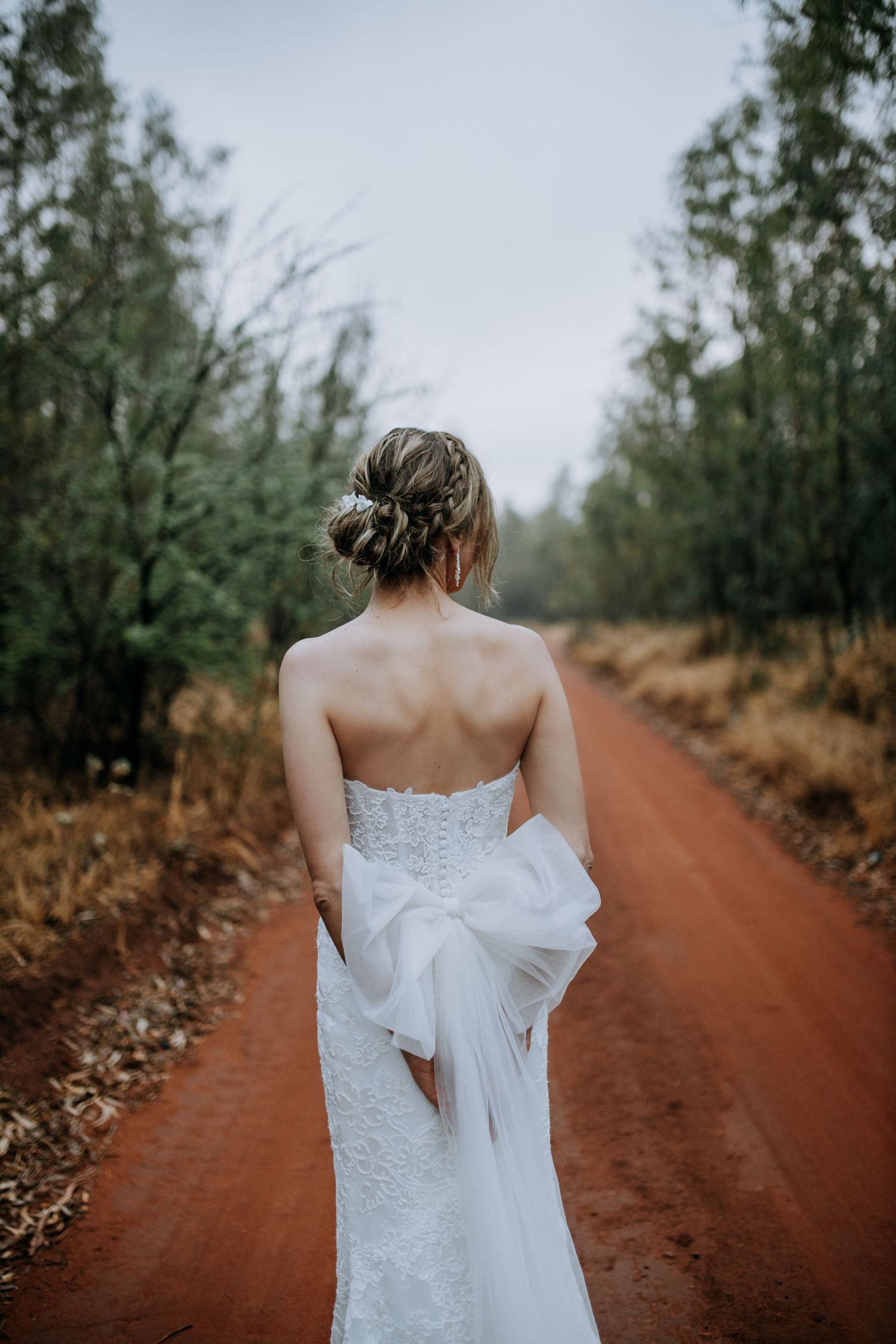 White wedding dress photographed from behind on a rustic red dirt road with trees in the background.