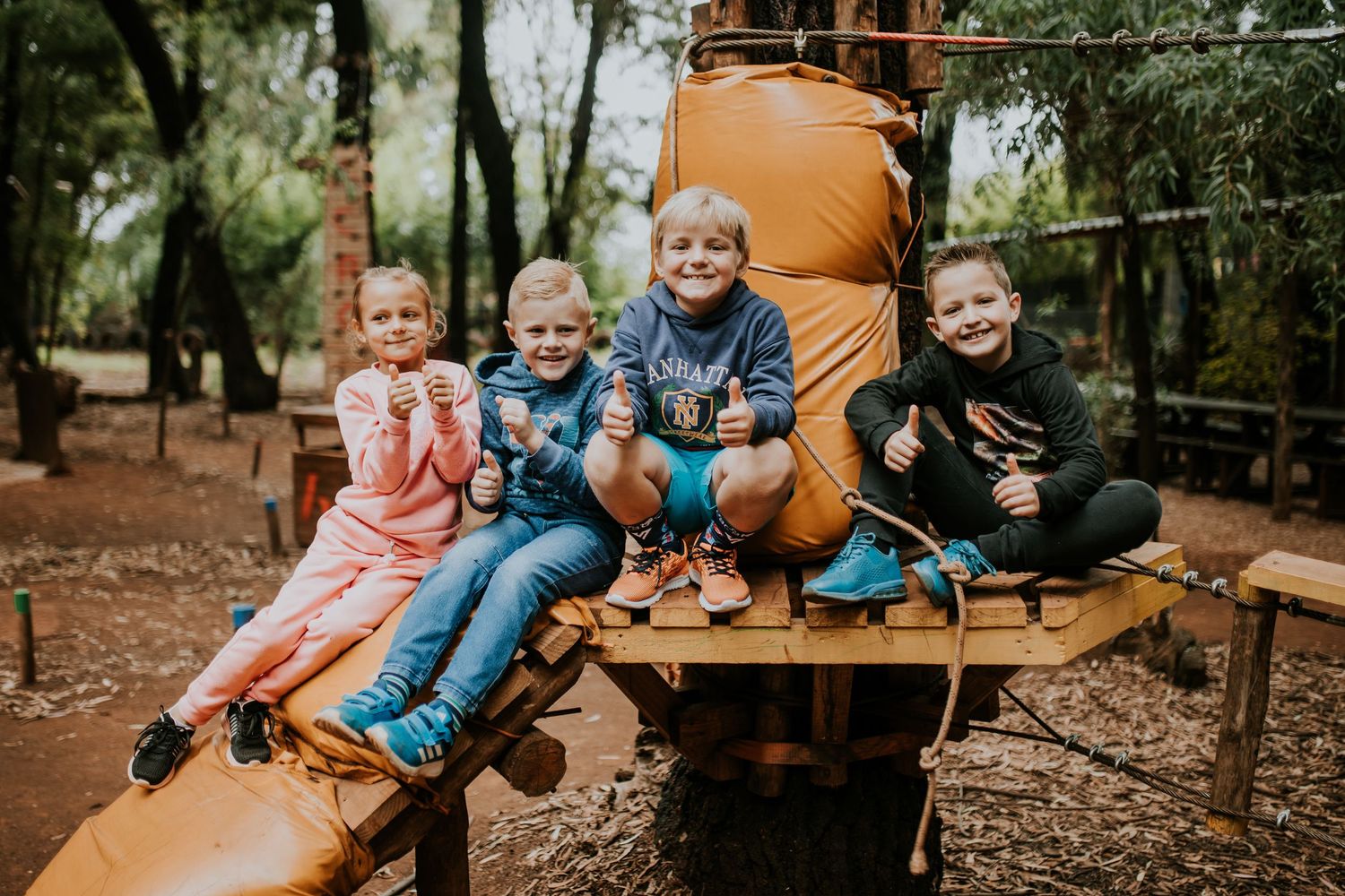 Children swing from a play rope at an outdoor adventure park with natural wood elements.