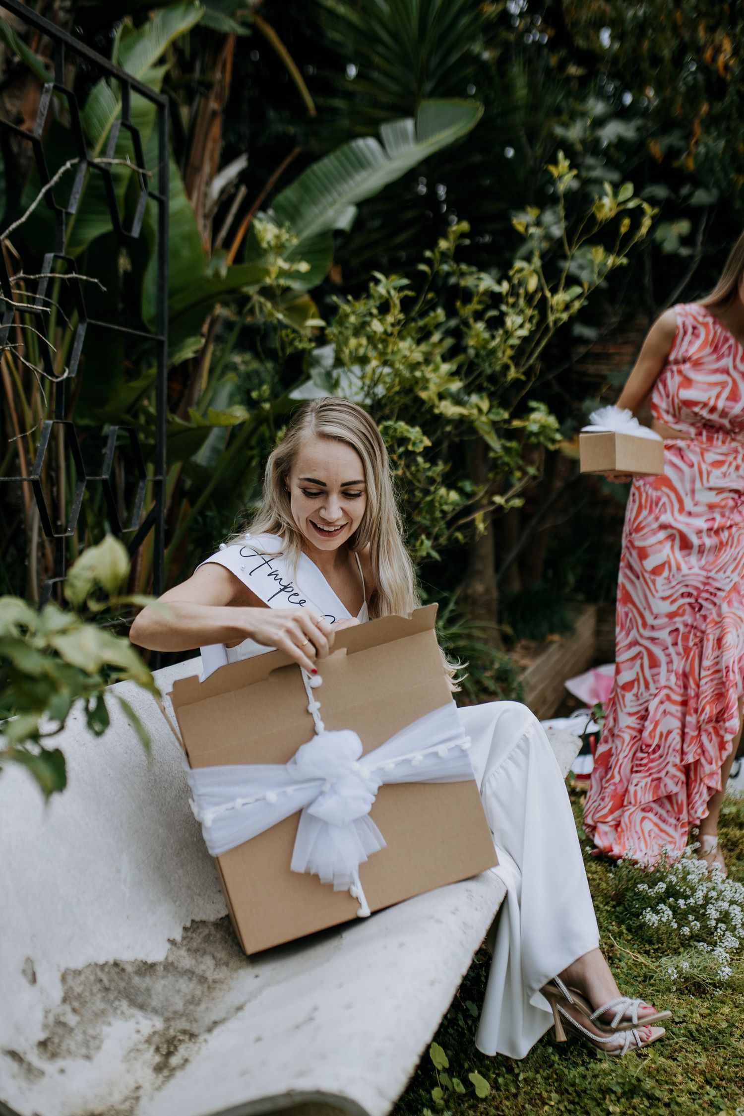 Person in a white gown sits in a garden setting opening a cardboard box containing white tissue paper.