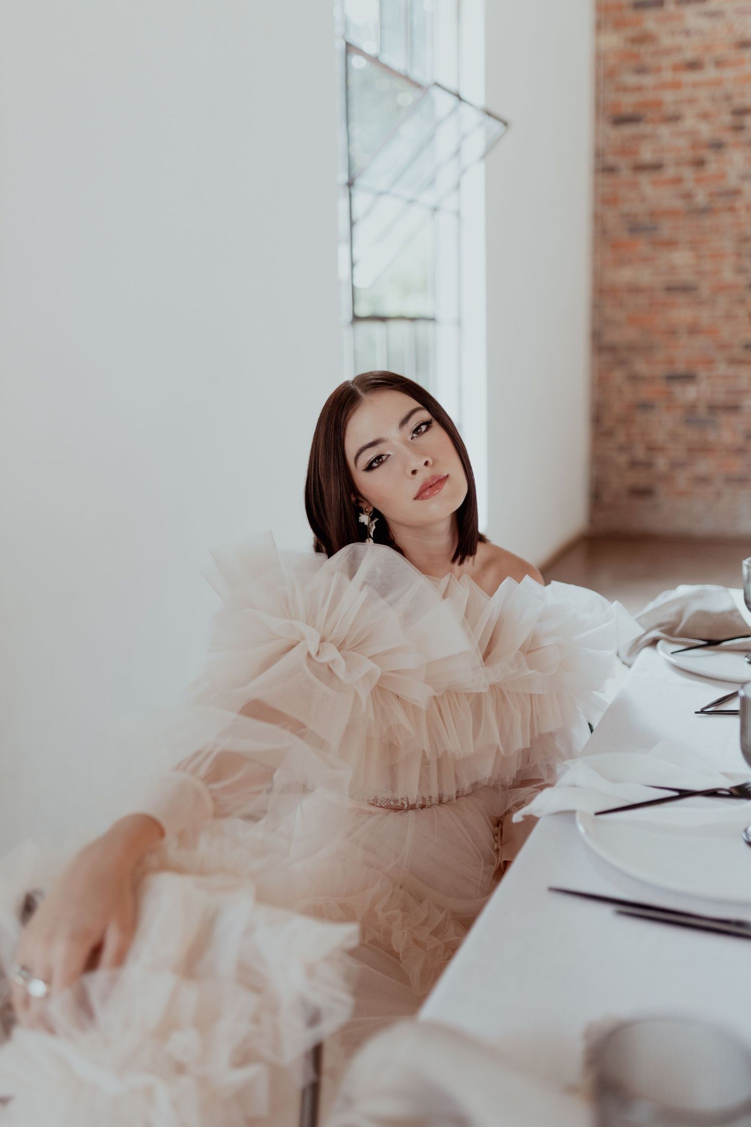 Fashion portrait against exposed brick wall featuring ethereal blush pink tulle dress in dreamy studio lighting.