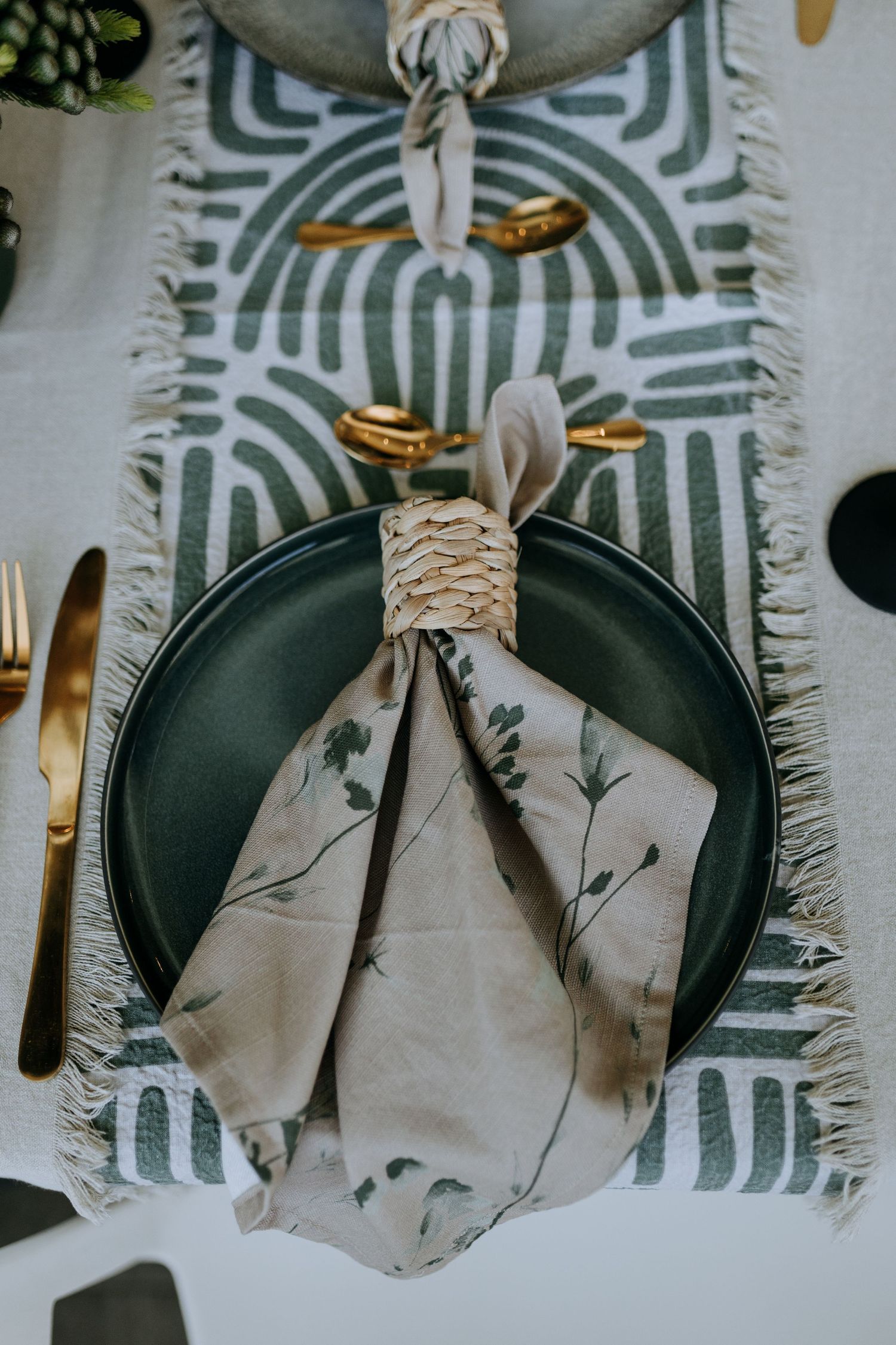 Elegant place setting with green plate, floral napkin tied with rope, and gold cutlery on geometric patterned table runner.