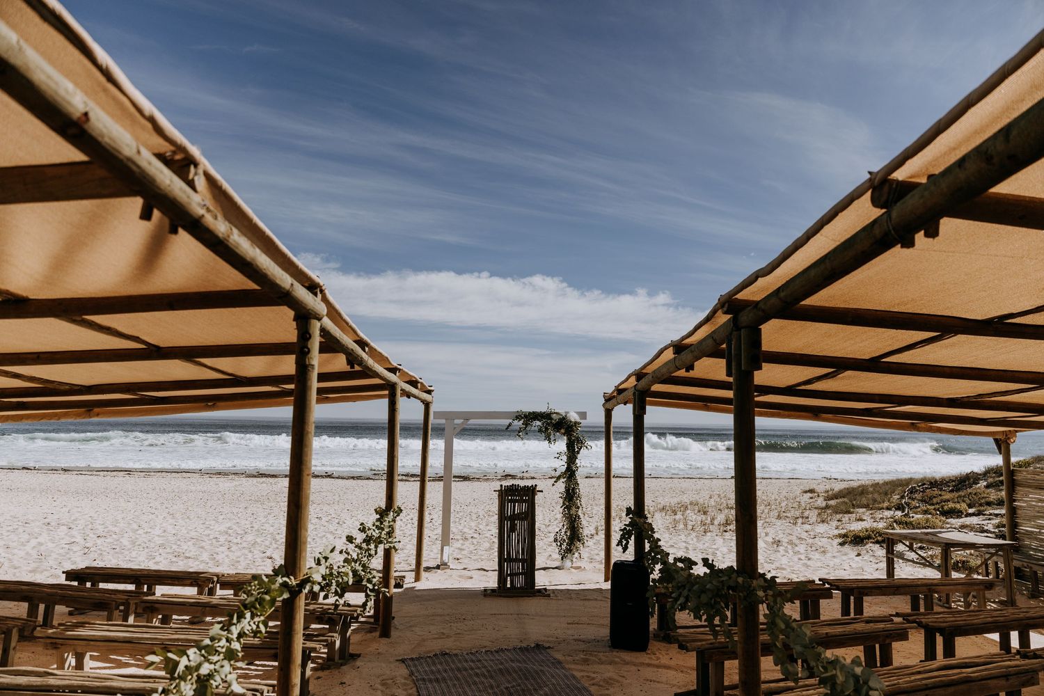 A beachfront restaurant deck with wooden pergolas and dining tables overlooking the ocean on a sunny day.