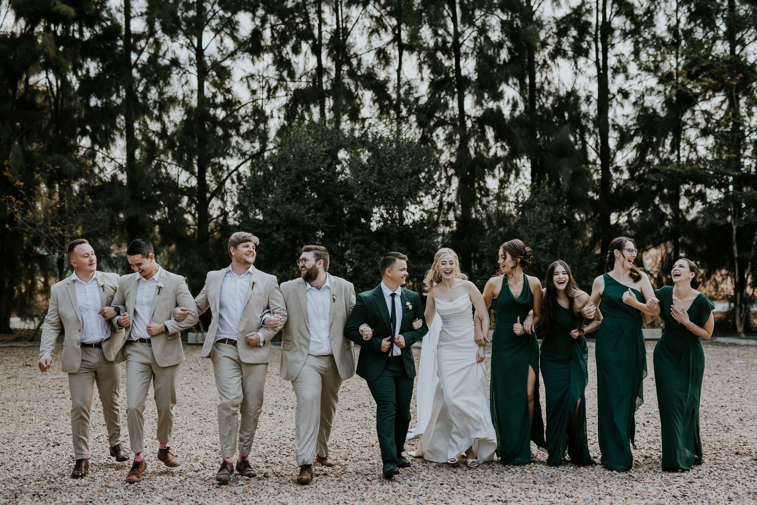 Looking down a line of bridesmaids and groomsmen standing outdoors against a wooded forest background as the sun sets.