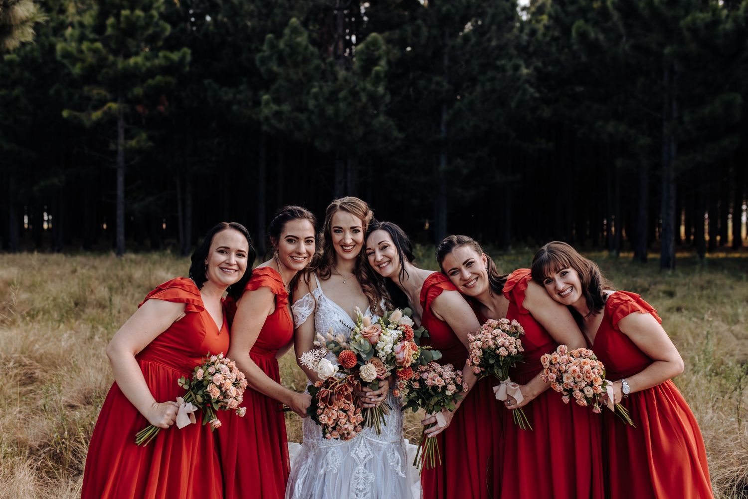 A wedding party wearing red bridesmaid dresses and a white wedding gown pose together in a rustic outdoor setting.