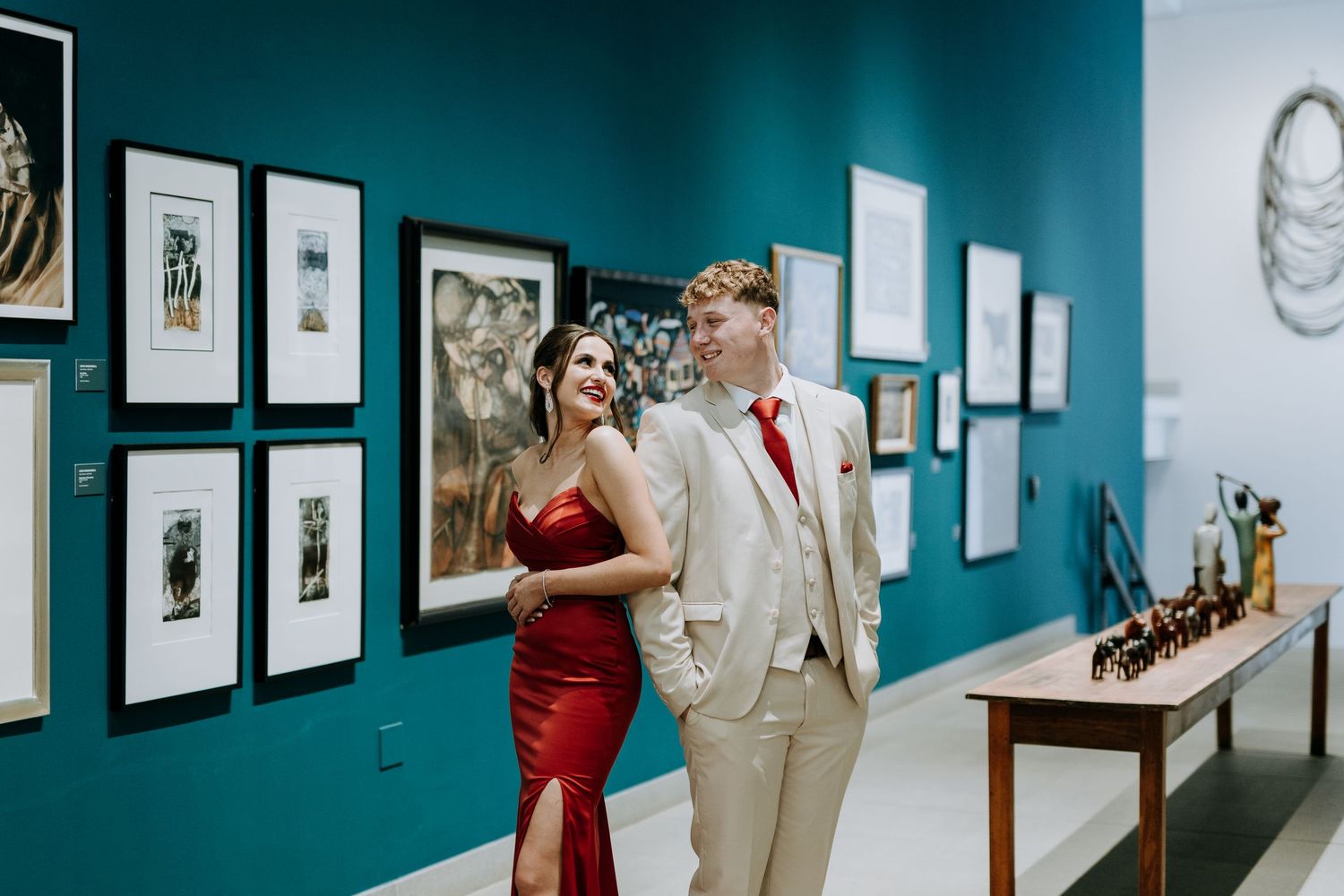 Art gallery visitors view framed artwork displayed on turquoise blue walls in a contemporary exhibition space.