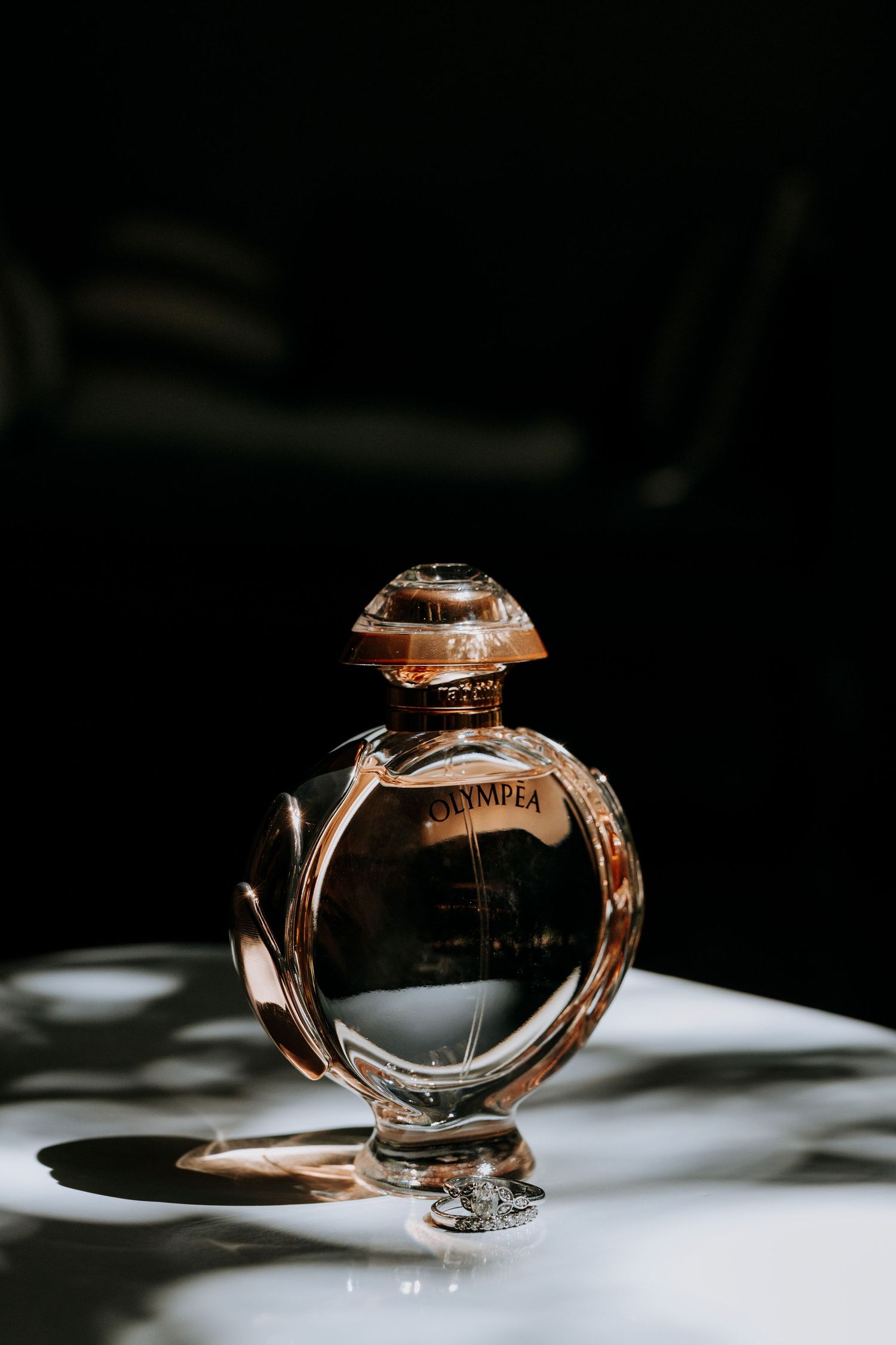 A rose gold perfume bottle photographed in dramatic lighting casting shadows on a white surface.