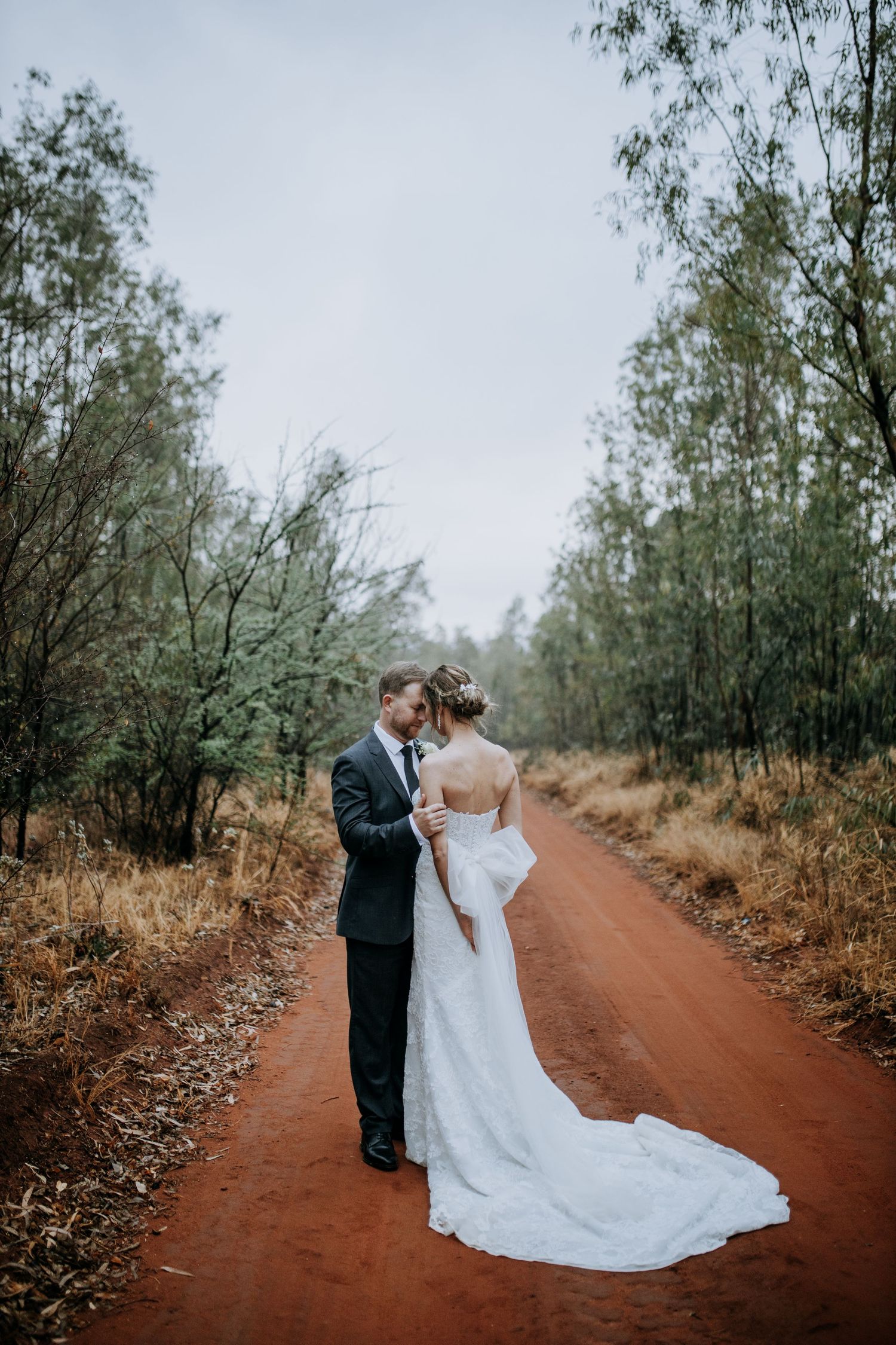 Wedding couple poses on red dirt road surrounded by eucalyptus trees.