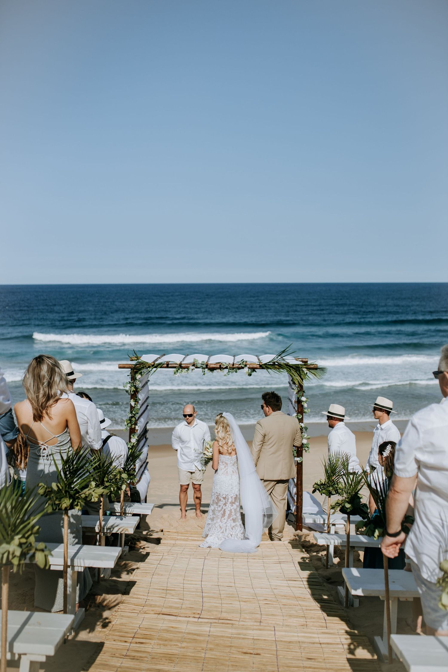 Wedding guests seated on wooden benches decorated with palm fronds line a beachfront aisle overlooking the ocean waves.
