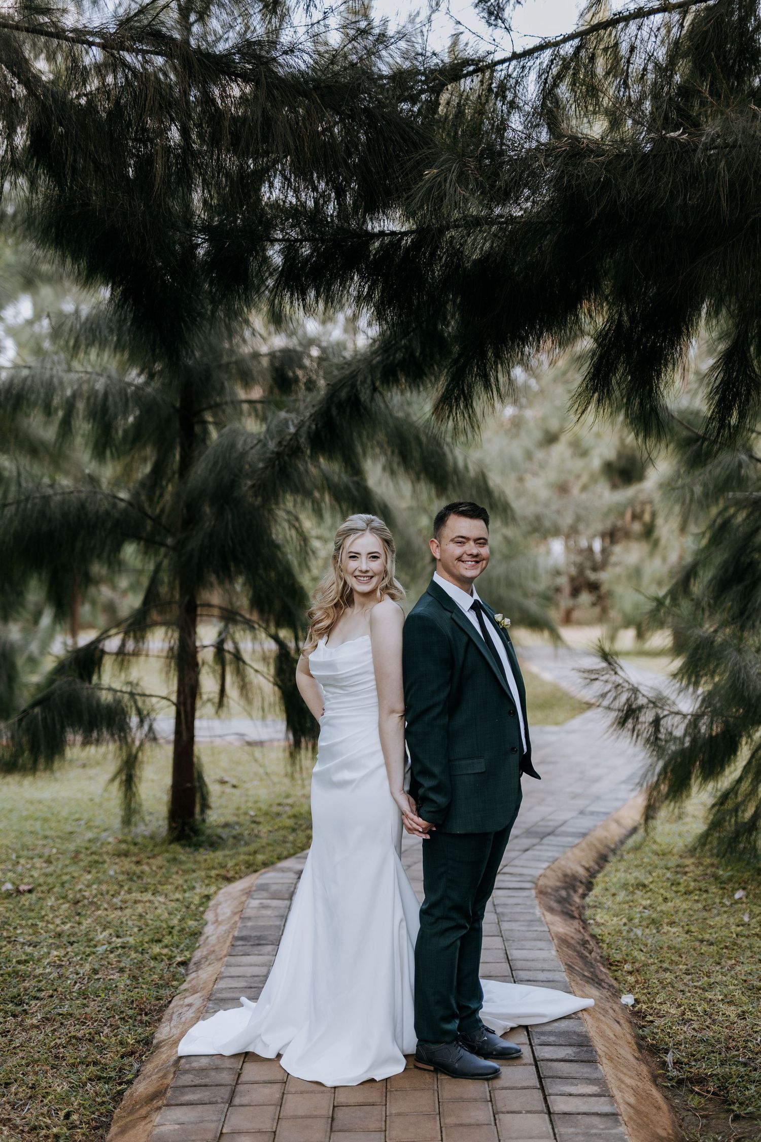 A newlywed couple shares playful moments on a stone path surrounded by evergreen trees in an outdoor garden setting.