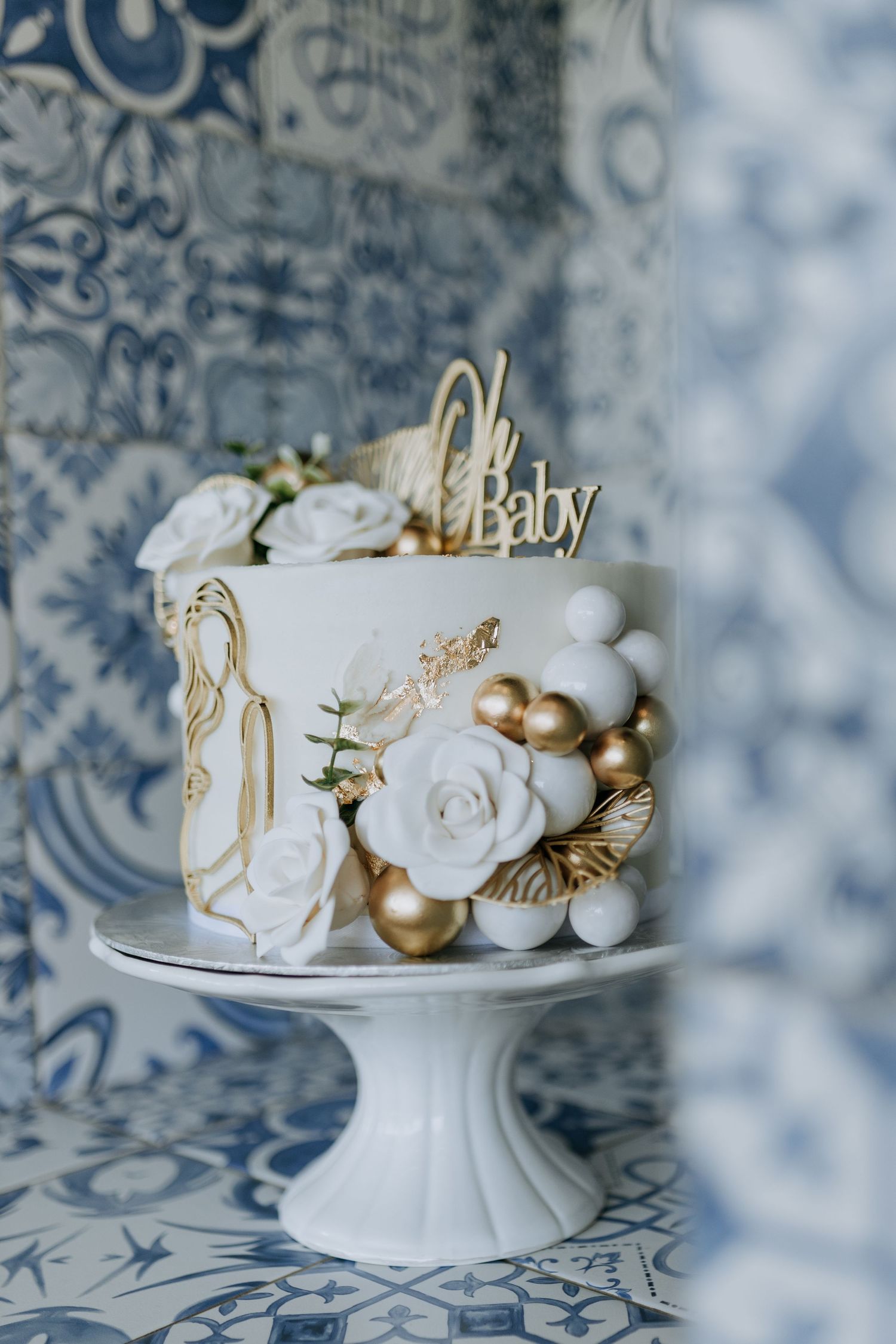 White ceramic decorative ornaments and golden baubles arranged on a cake stand against a blue and white patterned background.