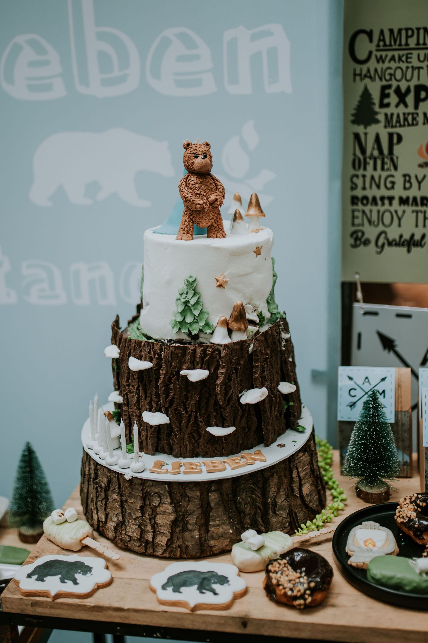 A rustic woodland themed cake decorated with tree bark texture, small bears, and miniature evergreen trees on a dessert table.